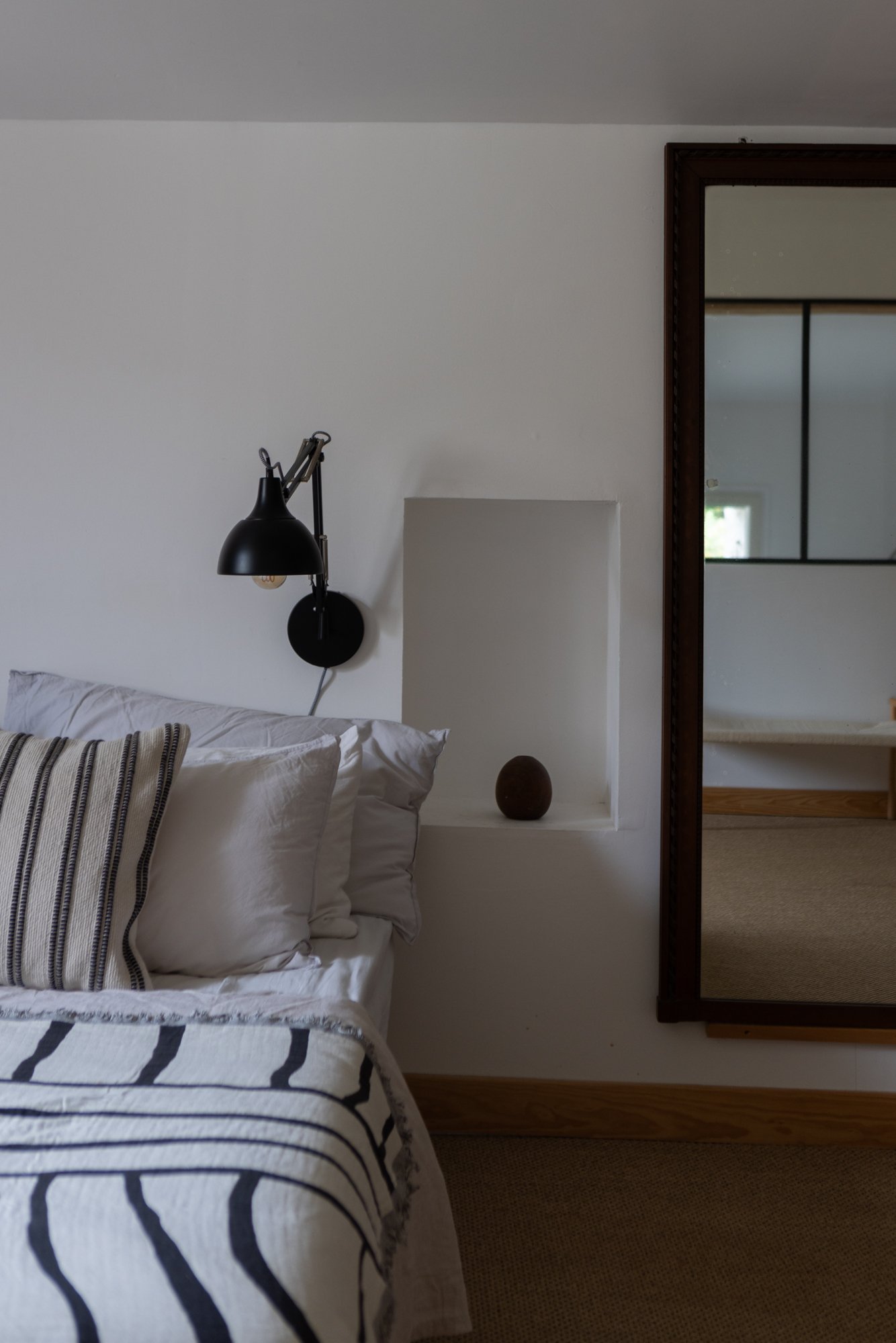 A cozy bedroom corner with a bed featuring white pillows and a black and white striped blanket, a wall-mounted black reading lamp, a small round dark brown vase on a built-in white shelf, a large rectangular mirror with a wooden frame, and a view int