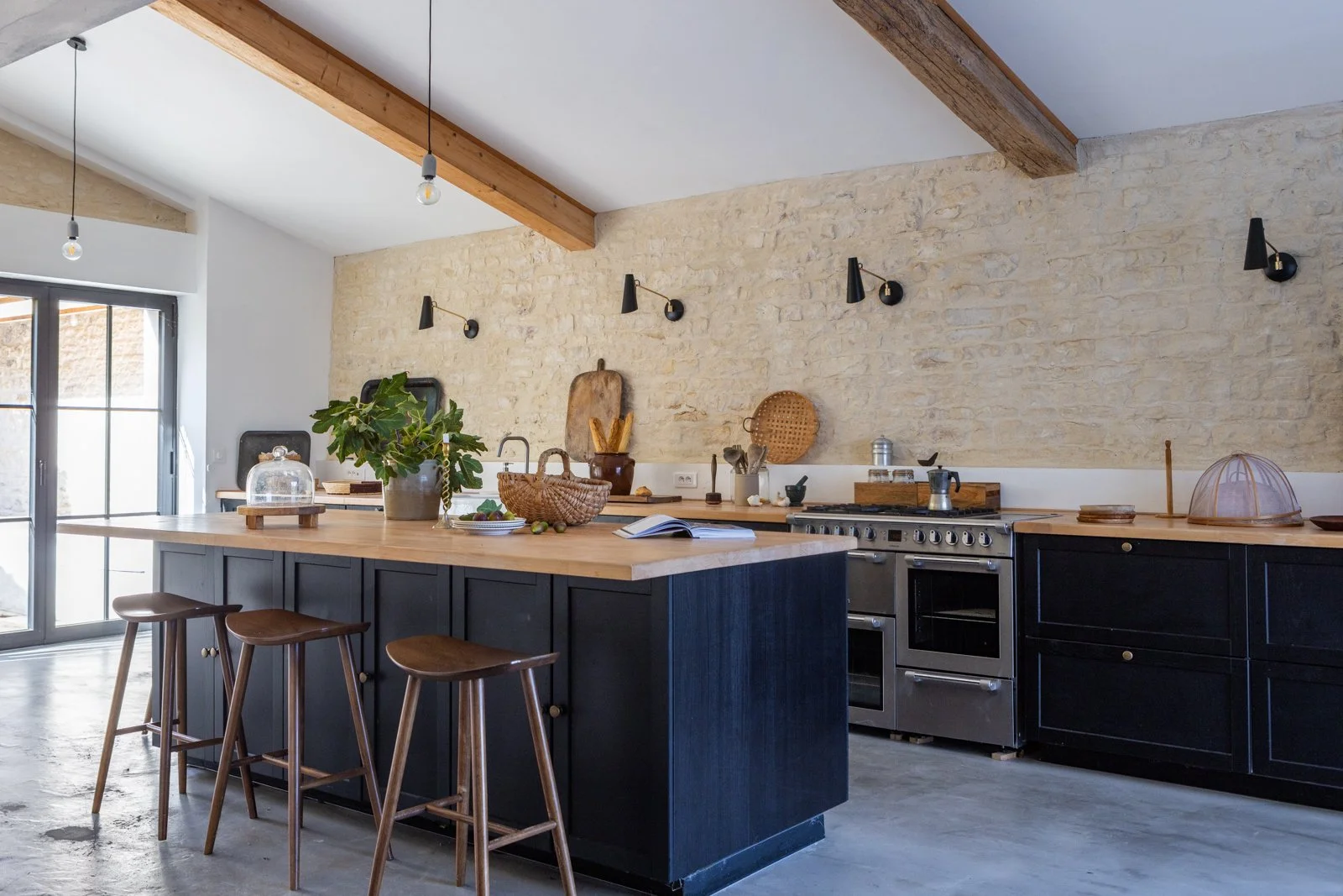 Modern kitchen with black cabinetry, wooden countertop, exposed brick wall, and wooden beams on ceiling. Decor includes plants, baskets, cutting boards, and kitchen utensils. Light fixtures hang from ceiling, and a sliding glass door to the outside i