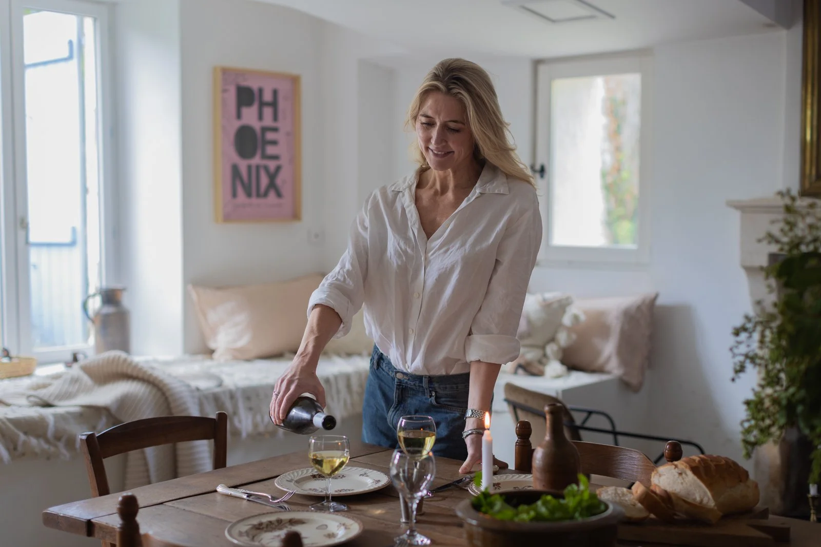 A woman with blond hair wearing a white shirt and jeans pouring white wine into glasses at a wooden dining table in a bright, cozy room.