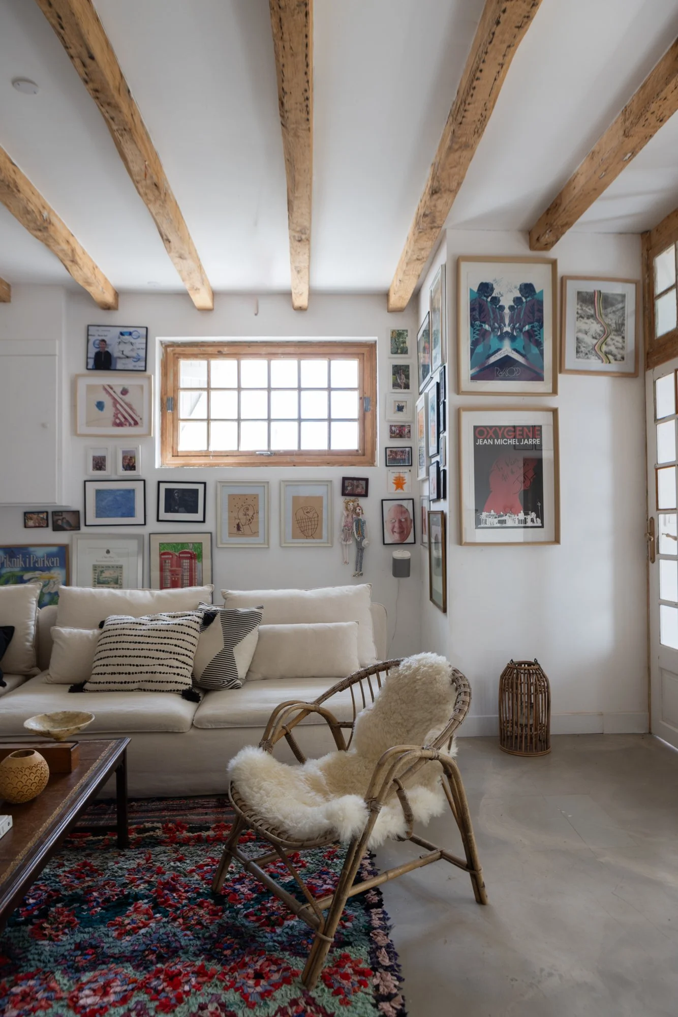 Cozy living room with cream couch, rattan chair with sheepskin cushion, wooden coffee table, colorful rug, gallery wall with framed art and photos, window with wooden trim, and natural light.
