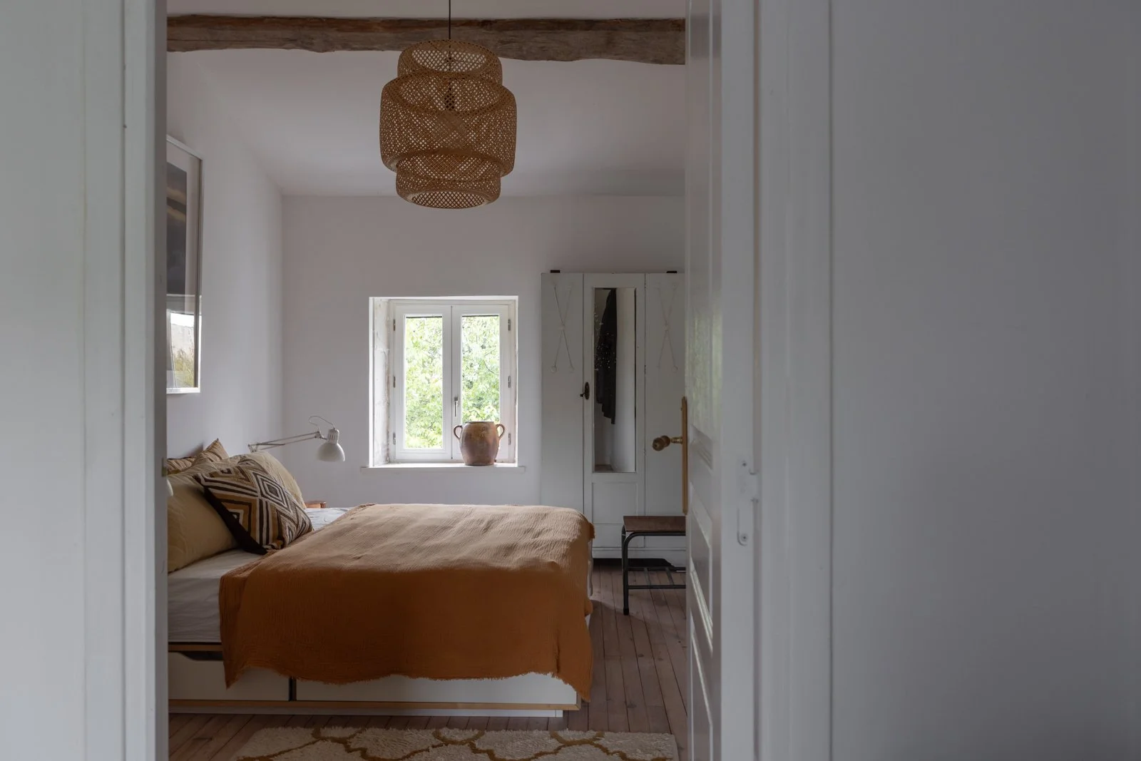 A cozy bedroom with a bed covered in a mustard-colored blanket, decorative pillows, a white wall-mounted lamp, a window with tree views, a white wardrobe, and a wicker chandelier hanging from the ceiling.