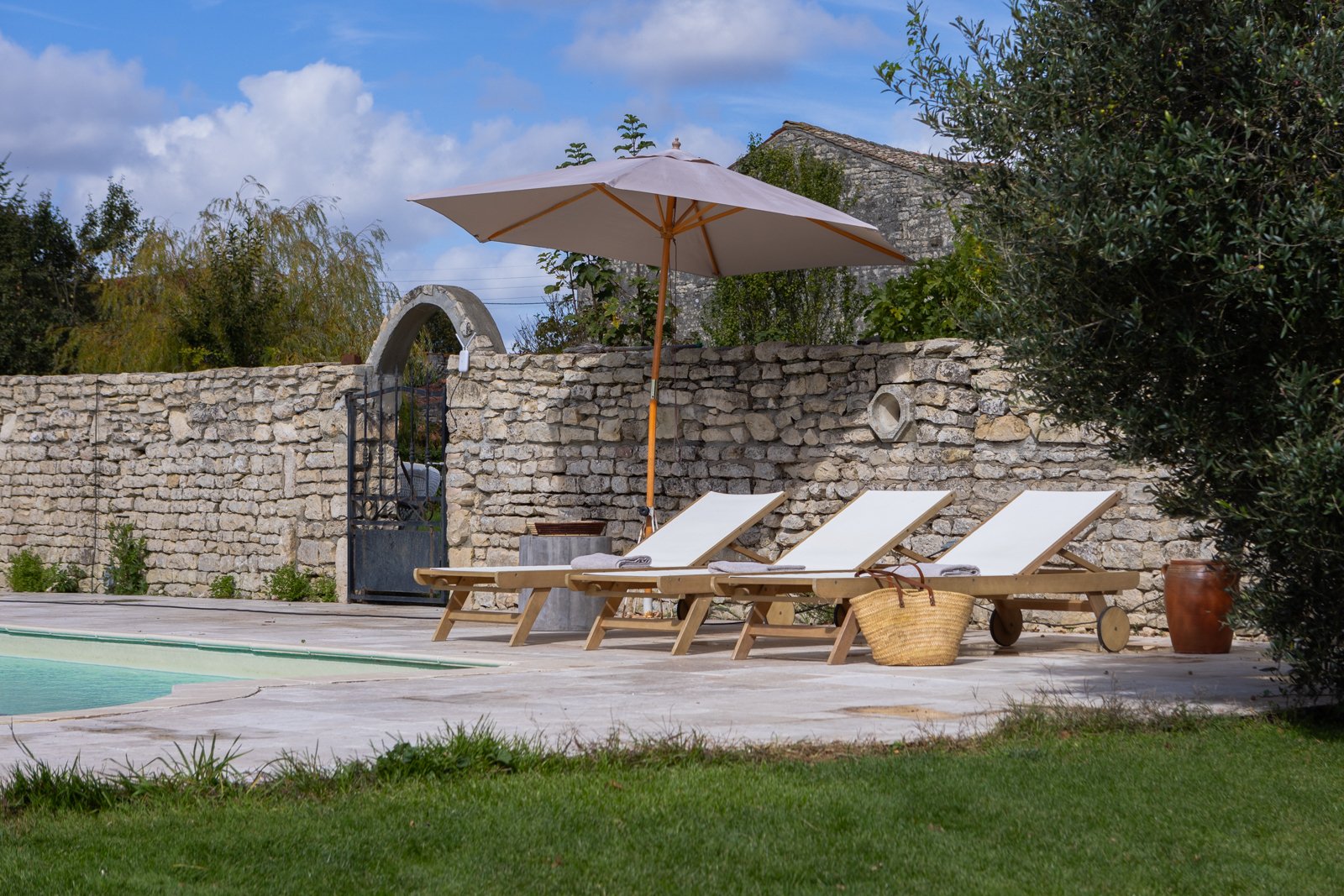 Three white lounge chairs with beige cushions and a wicker basket next to a swimming pool outside, with a stone wall, large tree, and partly cloudy sky in the background.