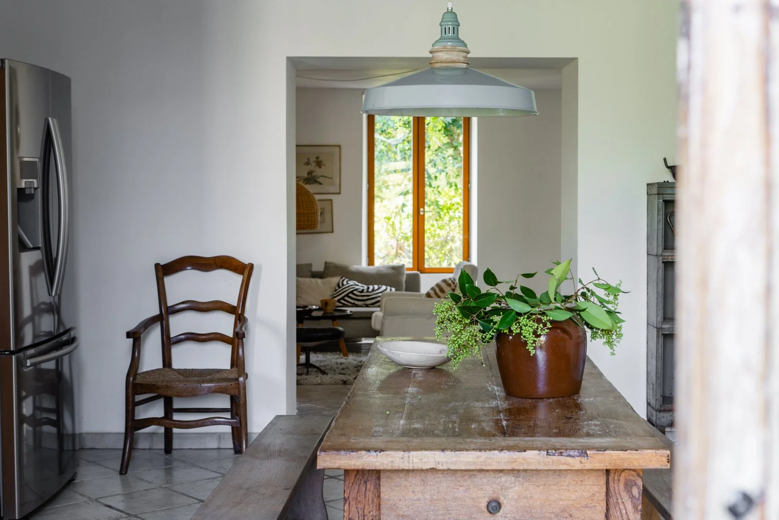 A cozy living room seen through a doorway from a rustic kitchen, with a wooden dining table with a green plant in a clay pot on top, and a window with a view of greenery.