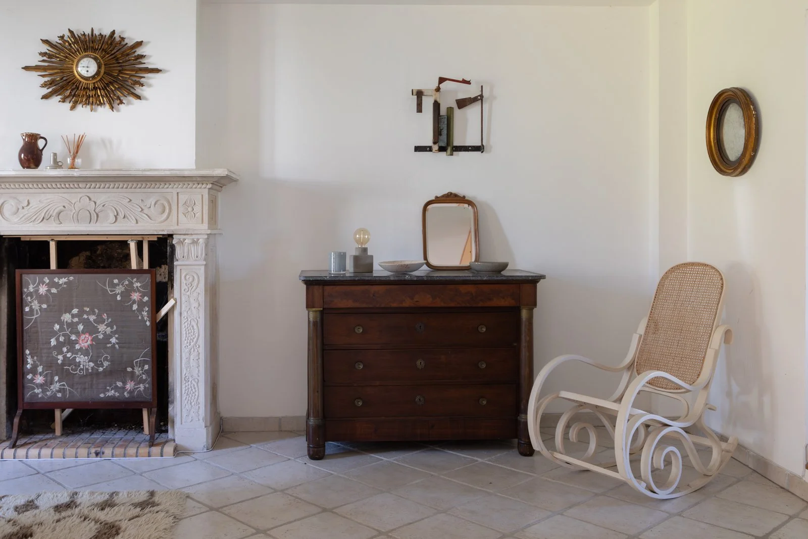 A cozy living room corner featuring a white fireplace with decorative carvings, an ornate gold sunburst clock above, a wooden dresser with a mirror and decorative objects on top, a vintage white rocking chair with a cane back, and a small round mirro