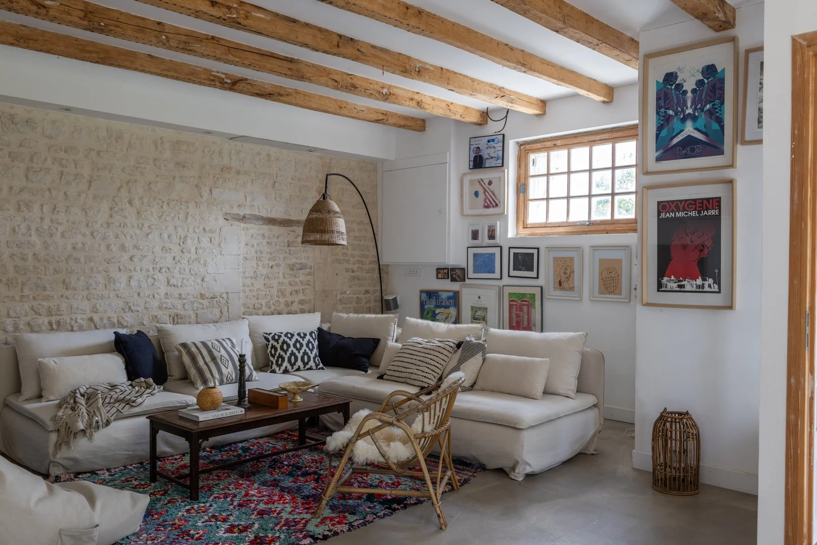 Living room with a white sectional sofa, various patterned pillows, a wooden coffee table, a wicker chair with sheepskin, and a colorful patterned rug. The room has an exposed stone wall, wooden beams on the ceiling, art prints on the wall, and a sma