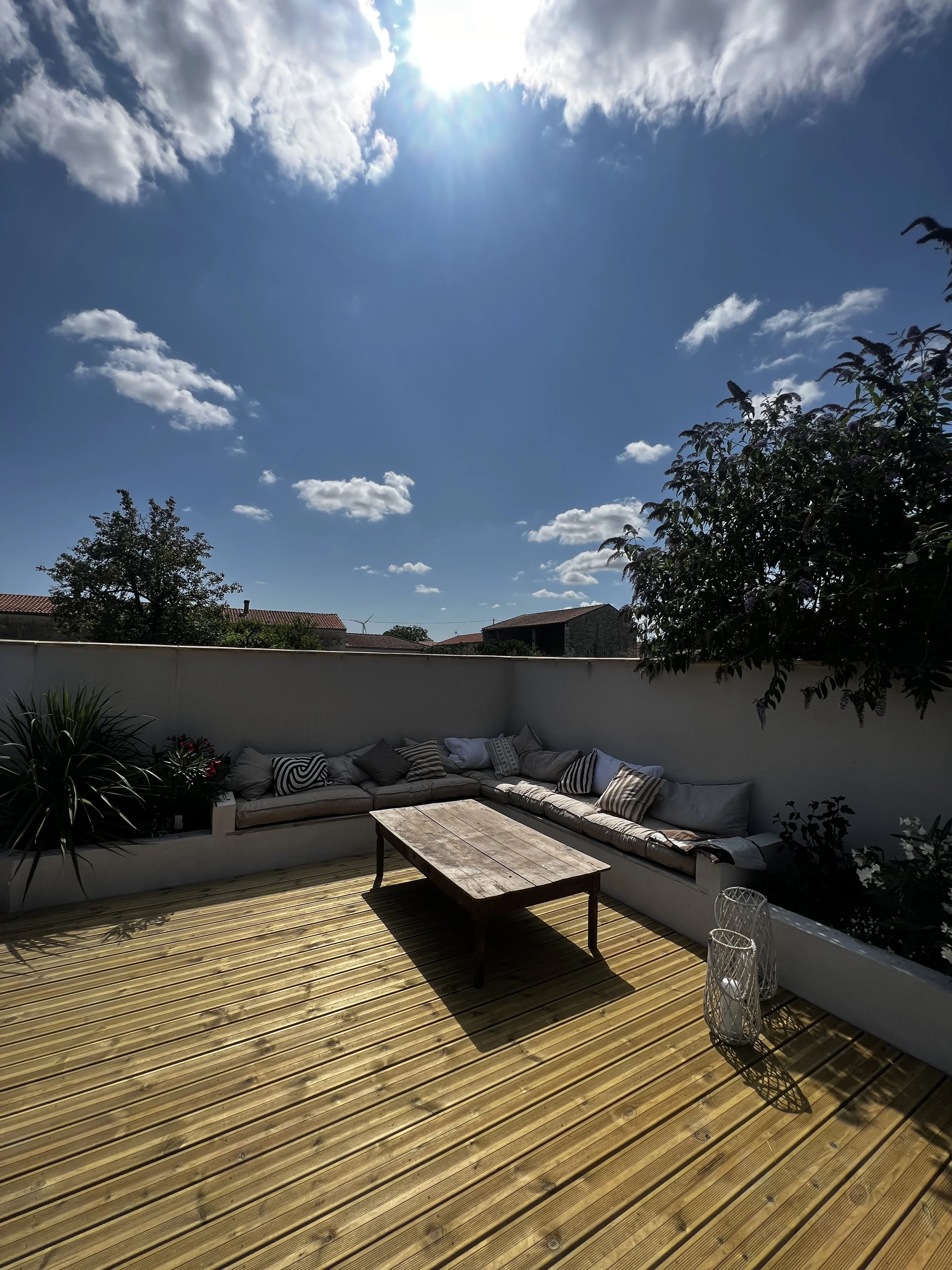 A sunny outdoor patio with wooden flooring, a long cushioned white sofa with various pillows, a wooden coffee table, and two decorative lanterns. The patio is bordered by a white wall with some plants and trees visible beyond, under a bright blue sky with scattered clouds.