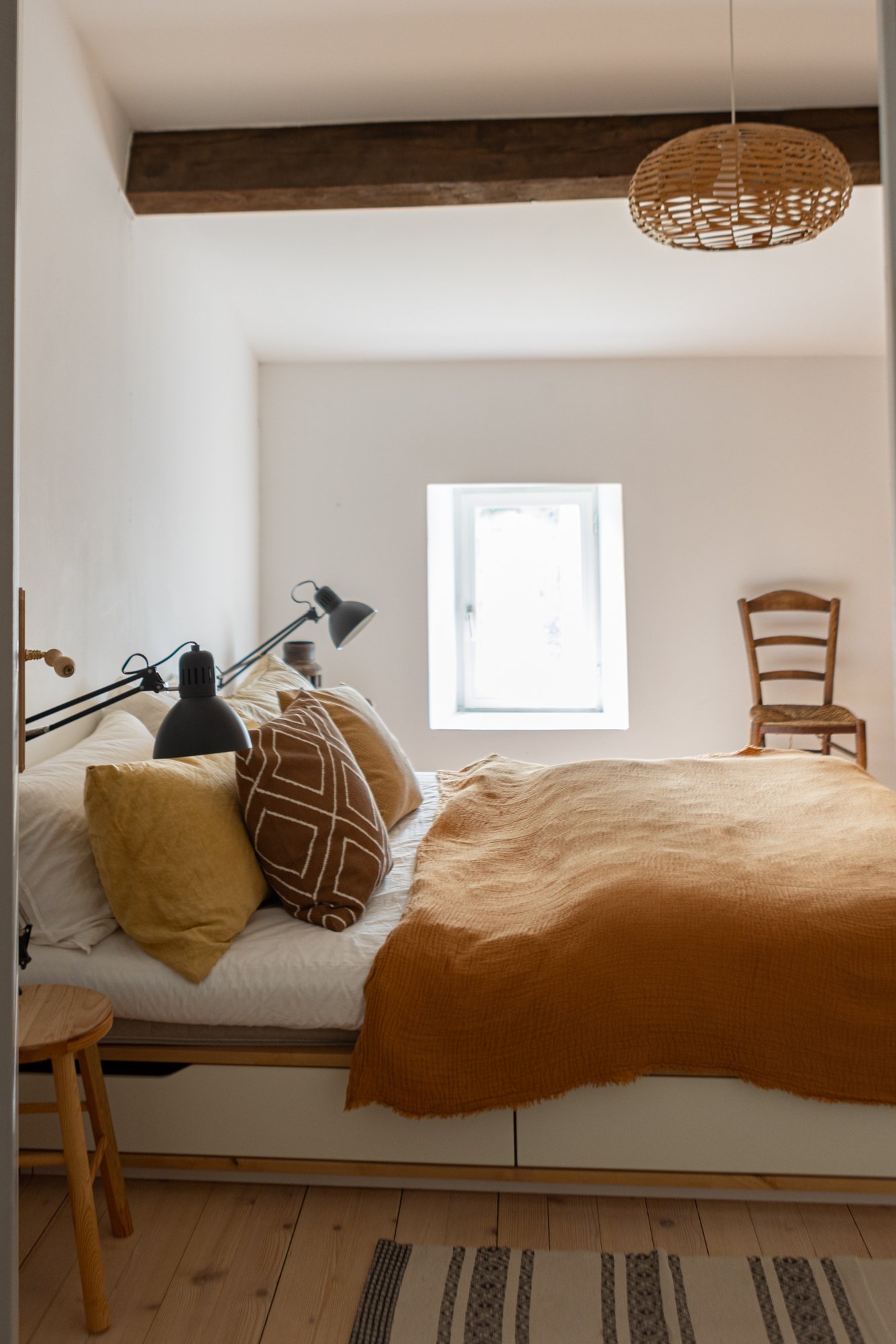 A cozy bedroom with a bed covered in rustic yellow and brown pillows and bedding, illuminated by natural light from a small window, with a wooden chair in the corner and a woven pendant light hanging from the ceiling.
