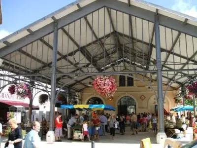 A busy outdoor market under a large metal canopy with hanging flower baskets, colorful umbrellas, and a crowd of people.
