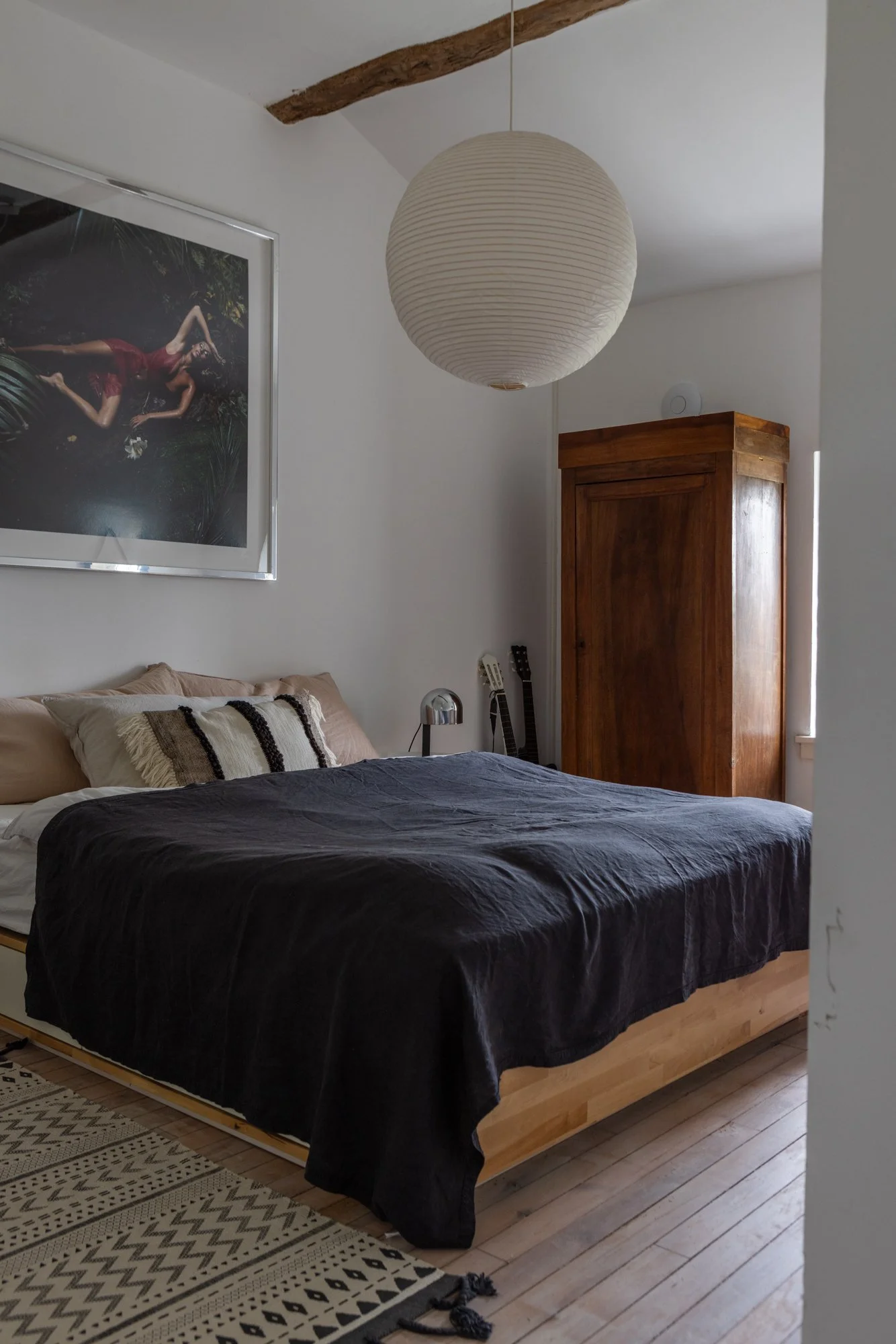 A bedroom with a bed that has beige pillows and a black bedspread, a large framed art print of a woman in a red dress on the wall, a wooden wardrobe, two guitars, a small silver lamp, a white lamp on the ceiling, a hanging paper lantern, and a patter