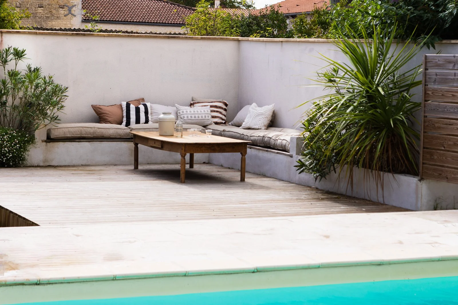 Outdoor patio with a pool, a wooden deck, a built-in gray concrete seating area with cushions and pillows, a wooden coffee table with decorative bottles, and lush green plants in a backyard setting.