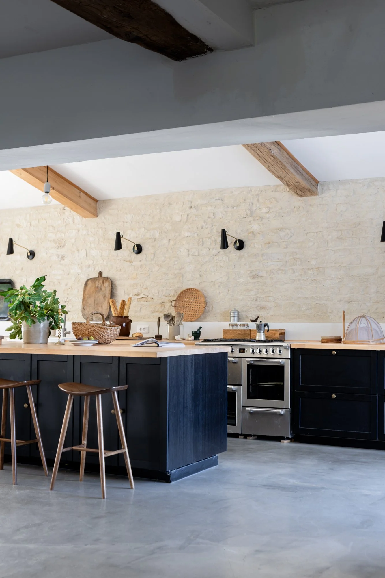 Modern kitchen with black cabinets, stainless steel stove, exposed brick wall, wooden island with two wooden stools, decorative plants, baskets, and wall-mounted black lights.