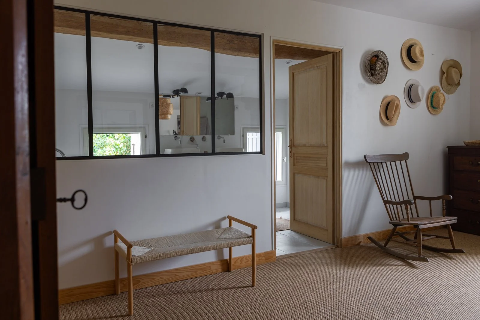 A room with a wooden bench and a rocking chair, a wall decorated with hats, an open doorway, and a large mirror reflecting a white wall and window.