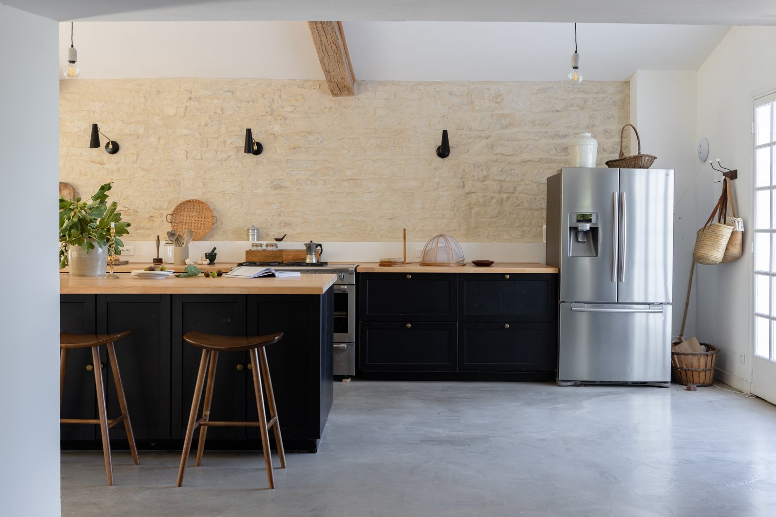 Modern kitchen with black cabinets, wood countertops, stainless steel refrigerator, and an exposed light bulb ceiling. Decor includes a wicker tray on the wall, a plant, and hanging bags.