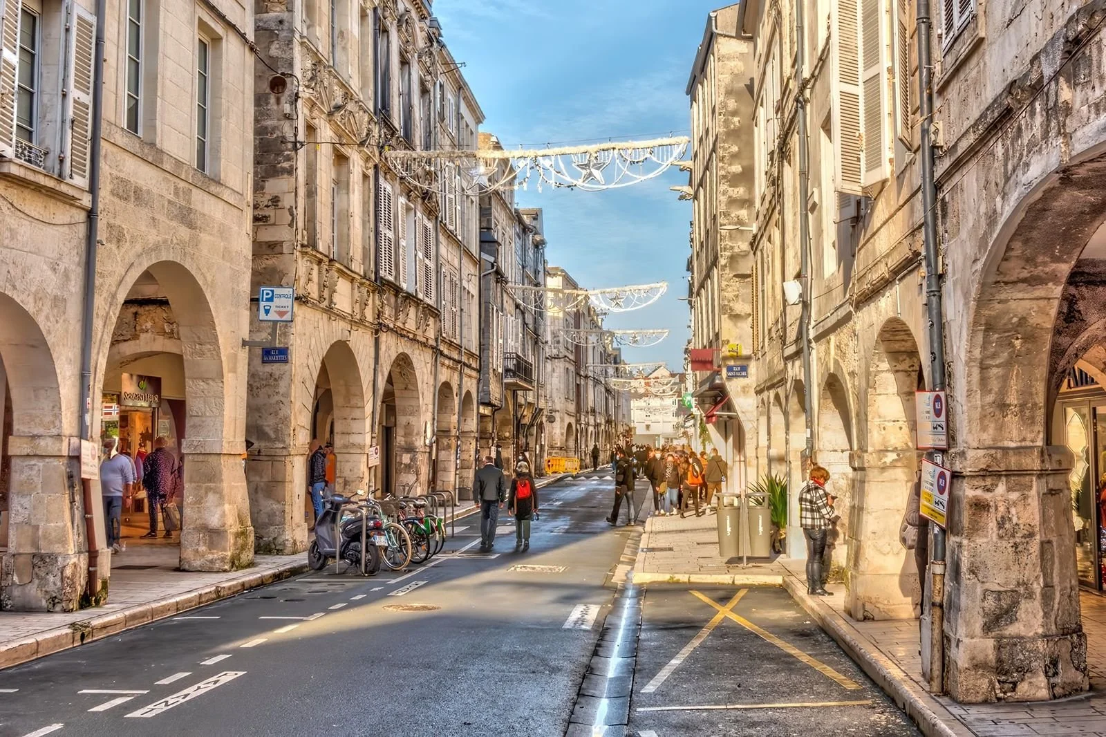 A busy street scene with historic stone buildings, arched walkways, and pedestrians walking along the sidewalks, with bicycles parked nearby and holiday decorations hanging across the street on a clear day.