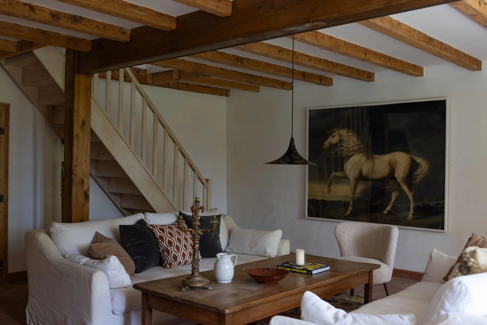 Living room with white sofas, wooden coffee table, large horse painting on the wall, wooden staircase, hanging lamp, and decorative items including a candlestick, a white pitcher, and books.