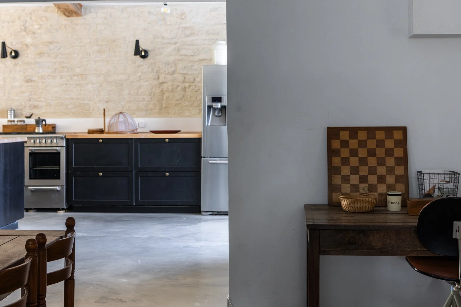 Interior view of a modern kitchen with blue cabinets and a stainless steel oven, with a partial view of a dining area featuring wooden chairs, and a separate desk area with a chessboard, basket, and jars.