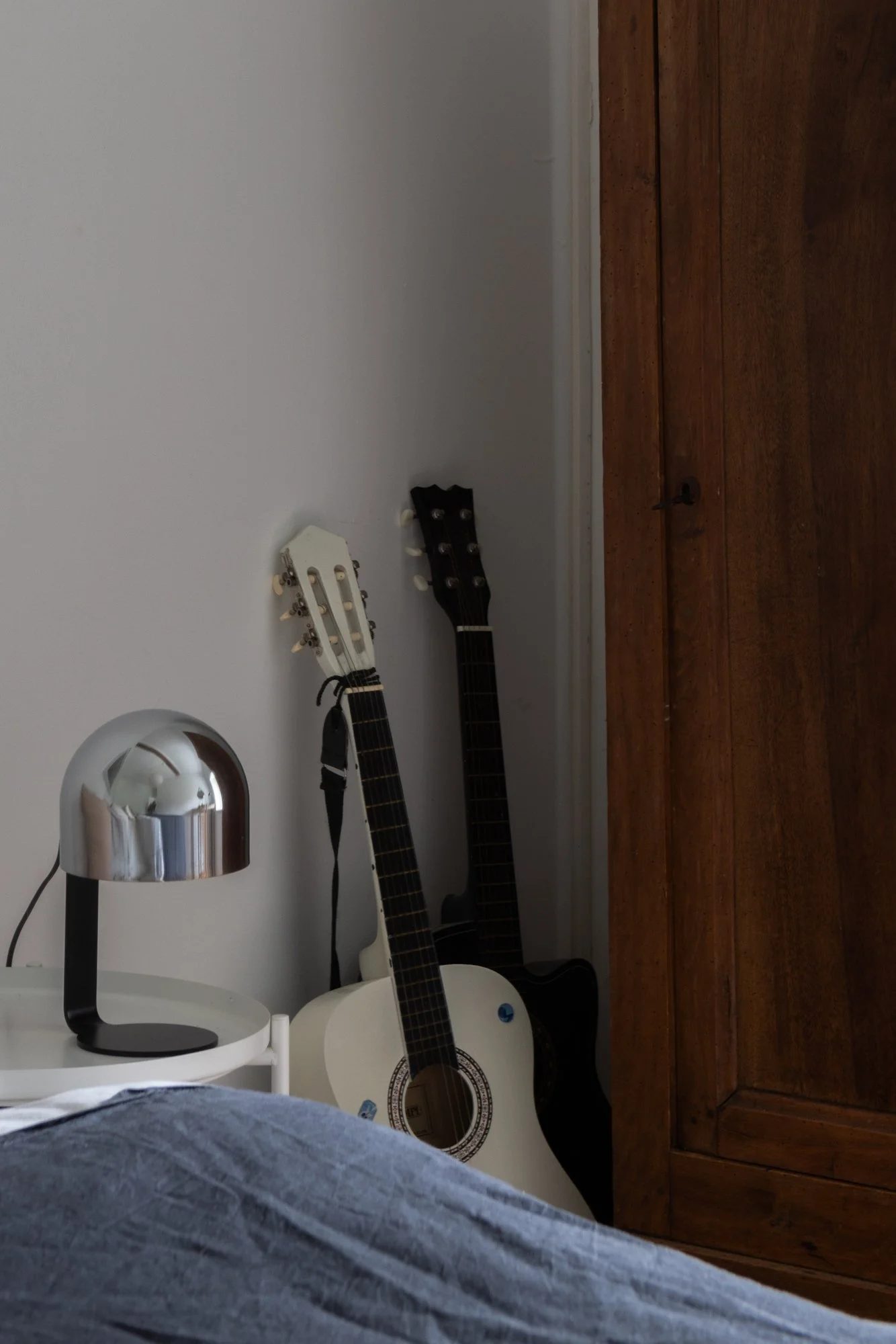 Two guitars, one white and one black, leaning against a white wall next to a dark wooden wardrobe, with a silver lamp on a white bedside table in the foreground.