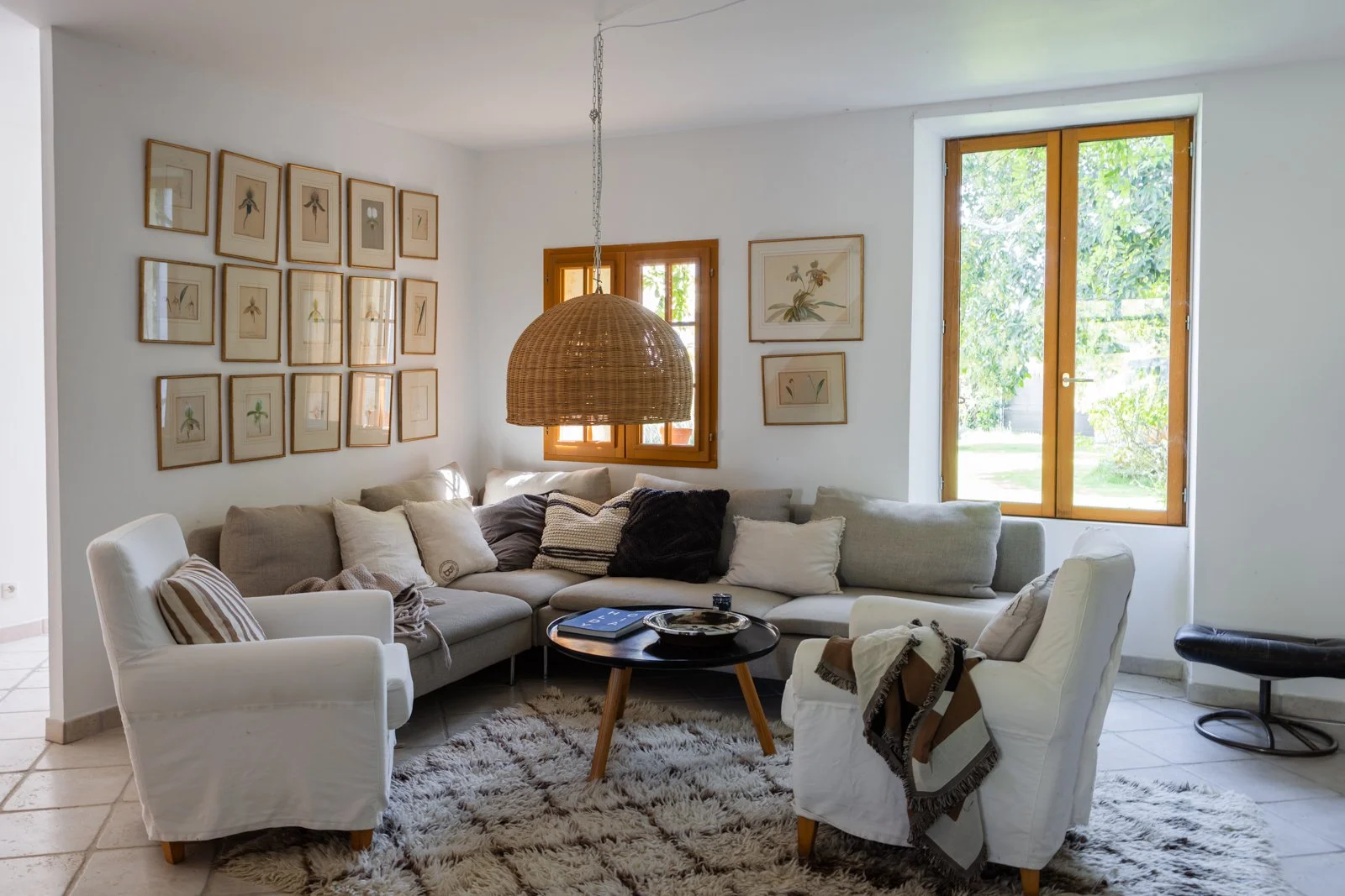 Living room with white walls, a large beige sectional sofa with throw pillows, two white armchairs, a black round coffee table, framed botanical art on the wall, wooden window frames, and a woven pendant lamp hanging from the ceiling.