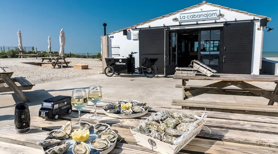 Outdoor seaside restaurant setup with oysters on the half shell, glasses of white wine, and a small black street food cart in front of a white building with black shutters and a sign that reads 'La Cabanajam'.