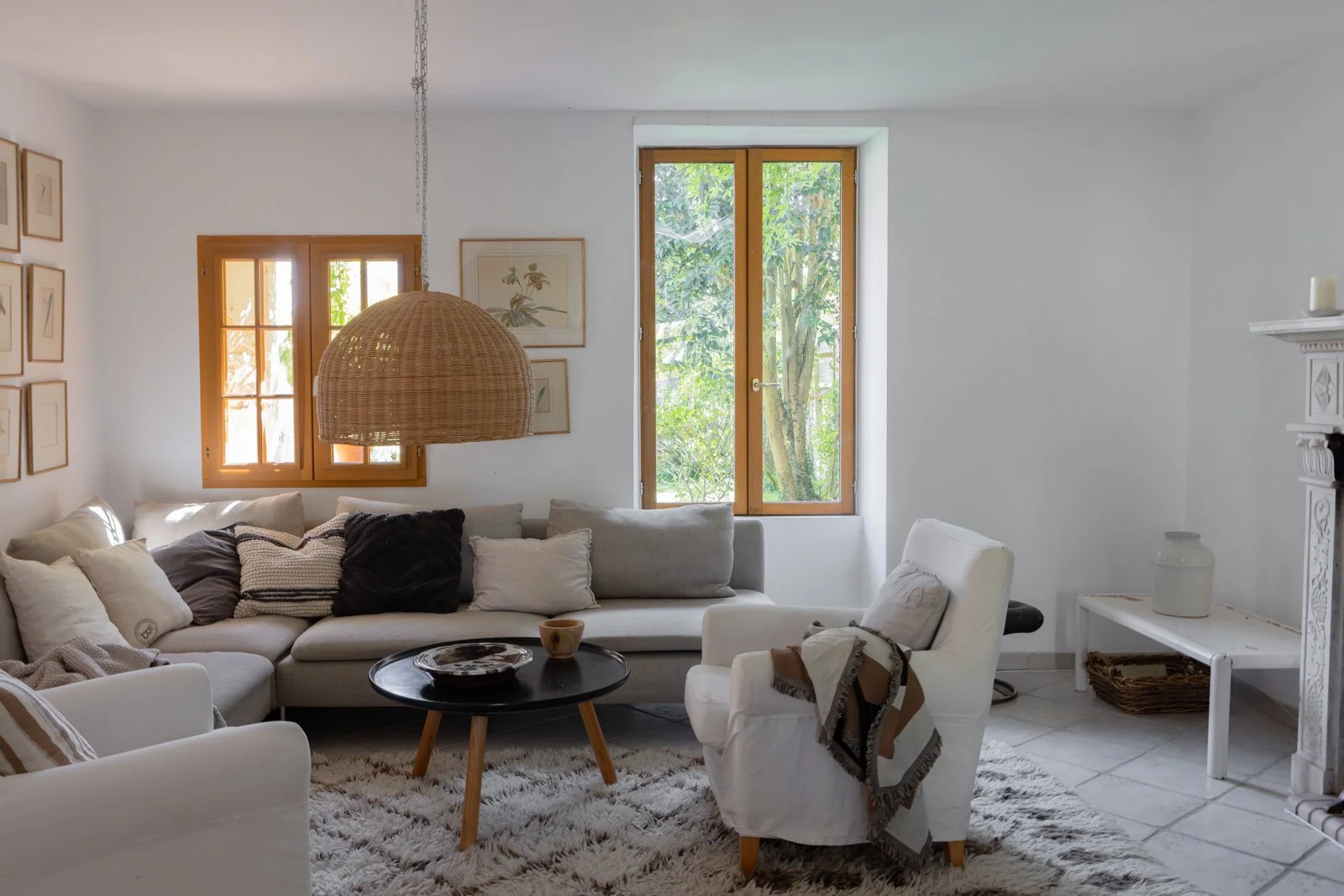 Bright living room with white walls, wooden-framed windows showing green foliage outside, a cozy beige sectional sofa with pillows, a white armchair with a blanket, a black round coffee table with a tray and cup, a woven pendant light, framed artwork