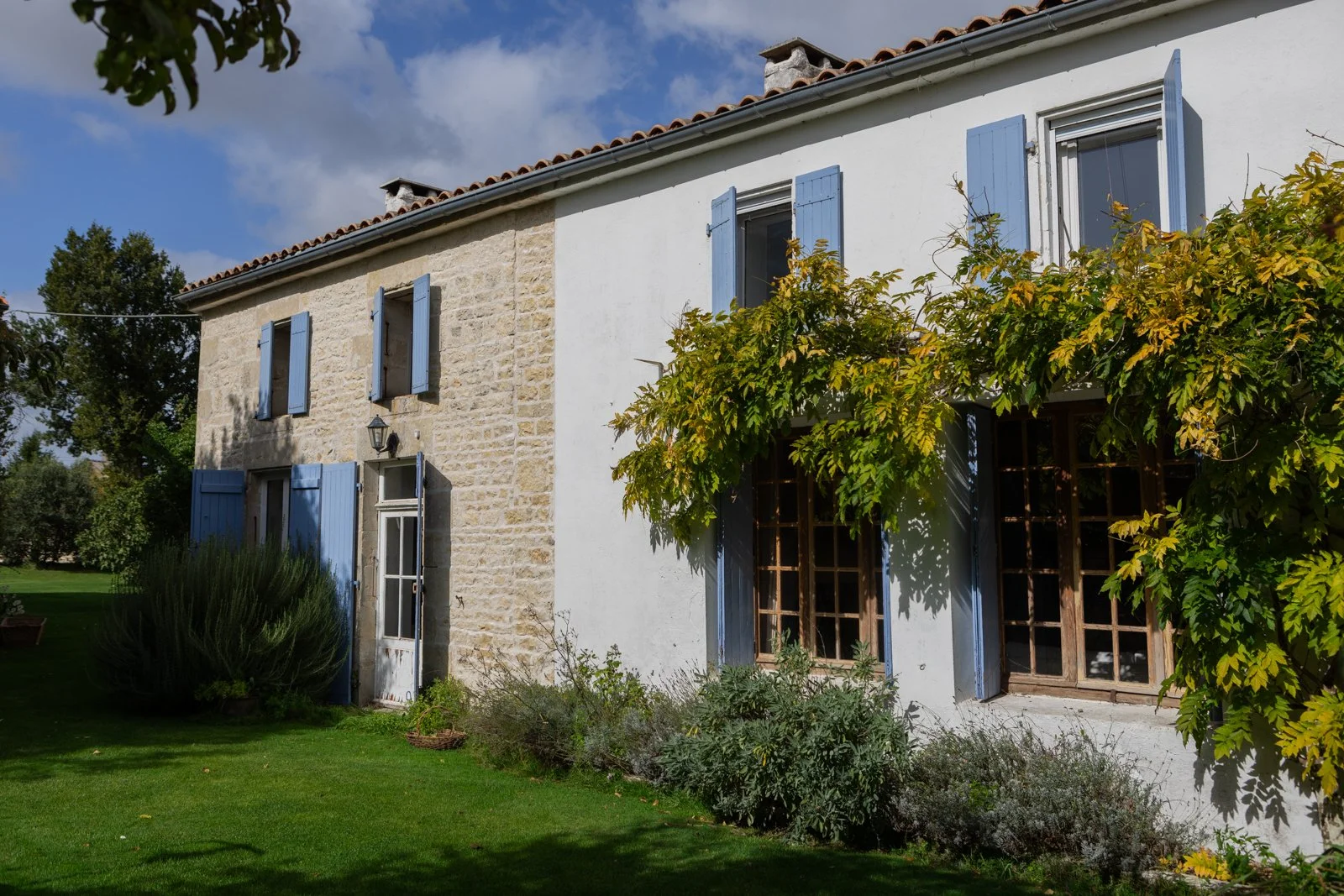 A two-story house with a stone and white stucco exterior, blue window shutters, and a garden with green grass and plants. The house has a red tiled roof and a tree with yellow and green leaves partially covering the front.