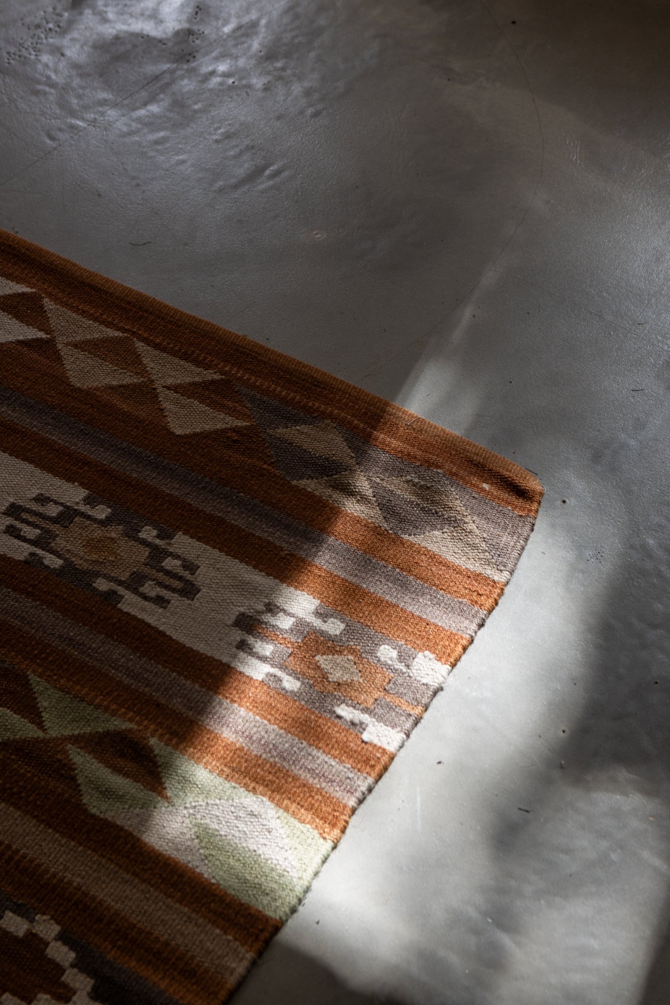 A corner of a patterned area rug on a concrete floor, with sunlight casting shadows.