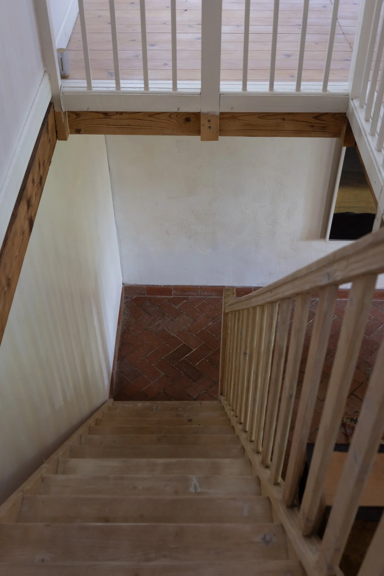 Top view of a staircase leading down to a brick floor, with a view of a wooden handrail and a walkway with a white railing at the top.