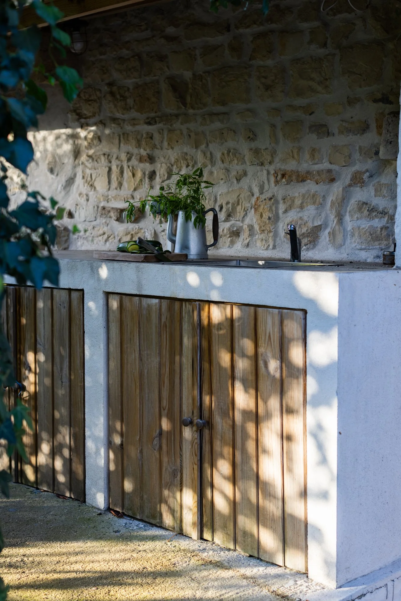 Outdoor kitchen with a stone wall and wooden cabinet doors, a black faucet, a potted plant in a metal watering can, and a cutting board with a knife and lemon on it, with dappled sunlight and shadow patterns.
