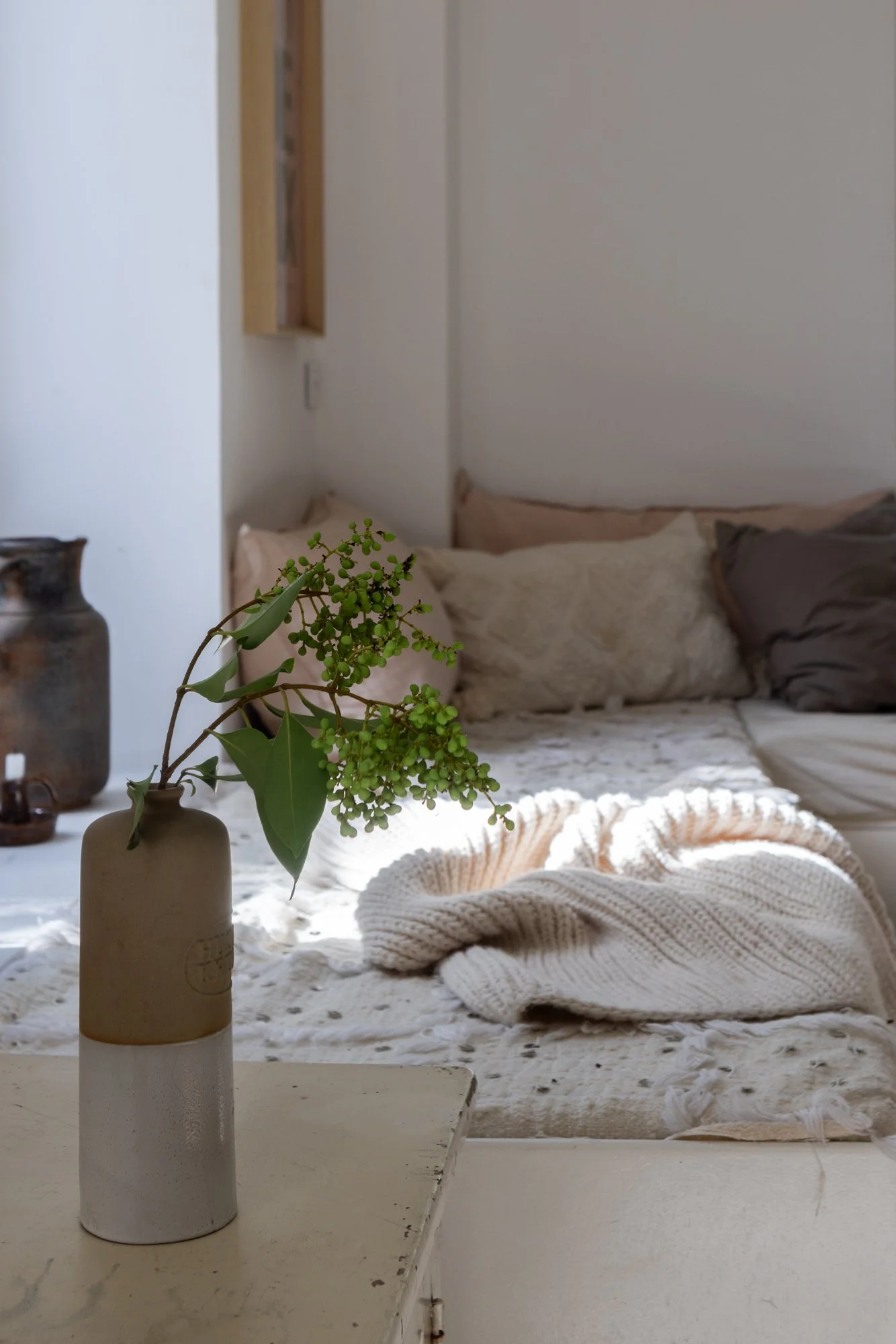 A beige and white ceramic vase with green leafy plant on a white table, with a bed featuring beige and grey pillows and a knitted blanket in the background.