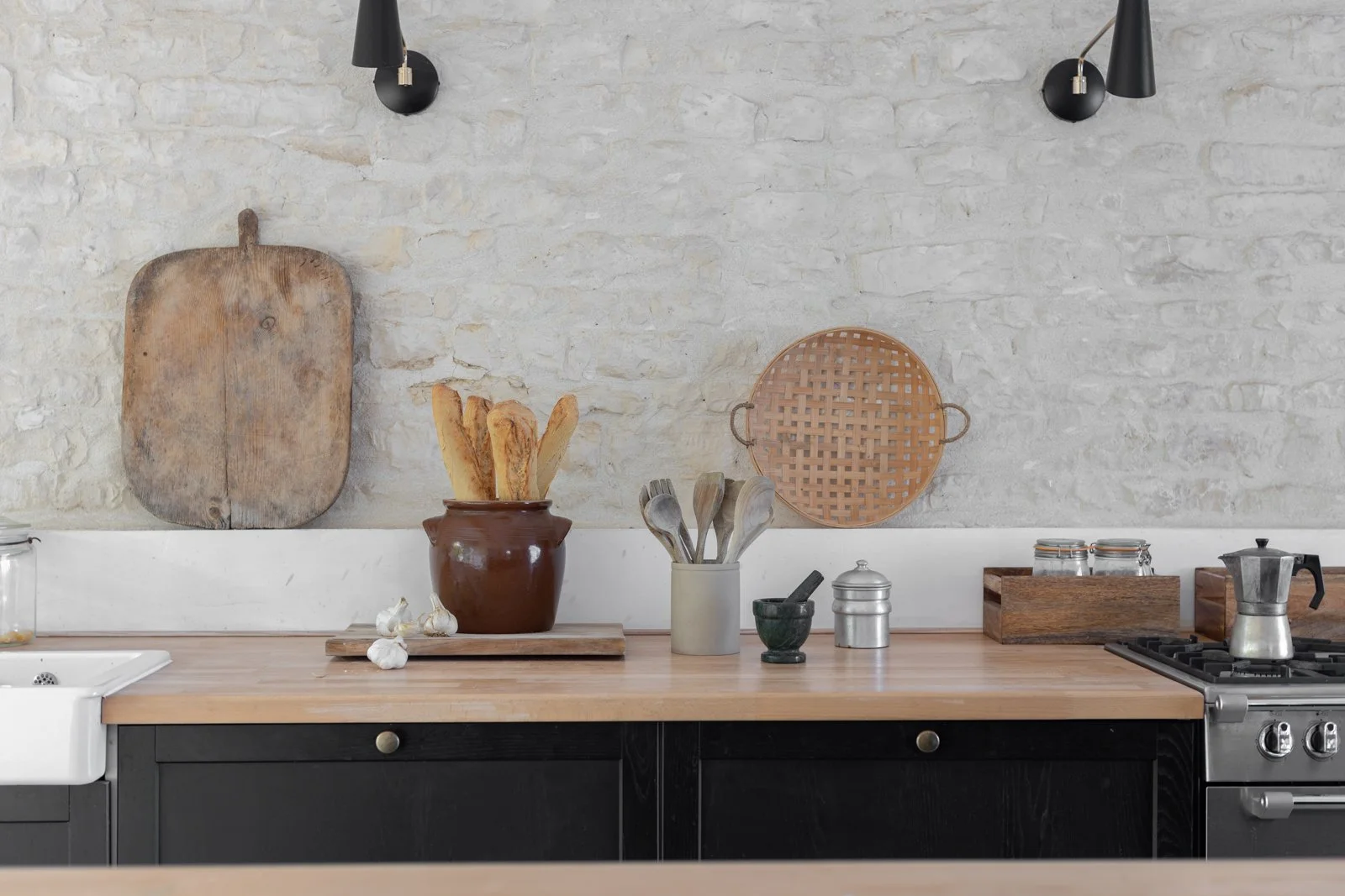 A rustic kitchen countertop with a white brick wall background. Items include a wooden cutting board, a pottery jar with bread, garlic, a utensil holder with wood spoons, a mortar and pestle, glass jars, a wooden tray, and a stove with a moka pot.