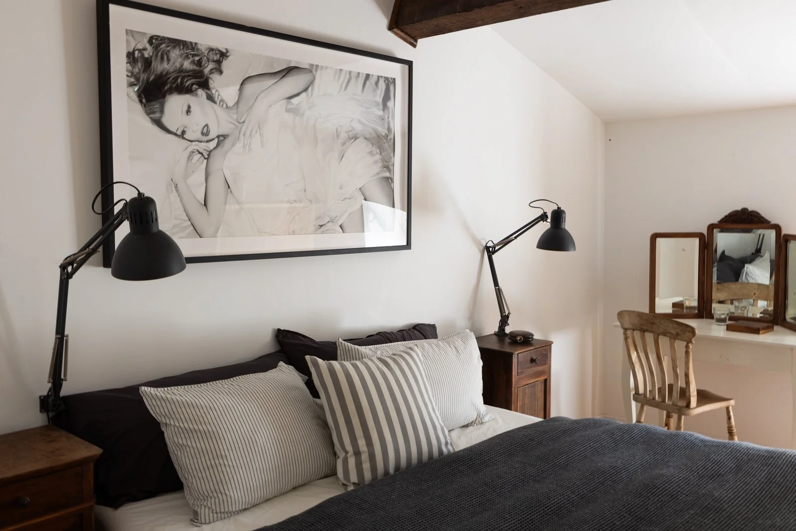 A cozy bedroom with a large black-and-white framed photograph of a woman lying down on the wall above the bed. The bed has striped pillows and a dark bedspread. There are two black adjustable wall-mounted lamps on either side of the bed. To the right