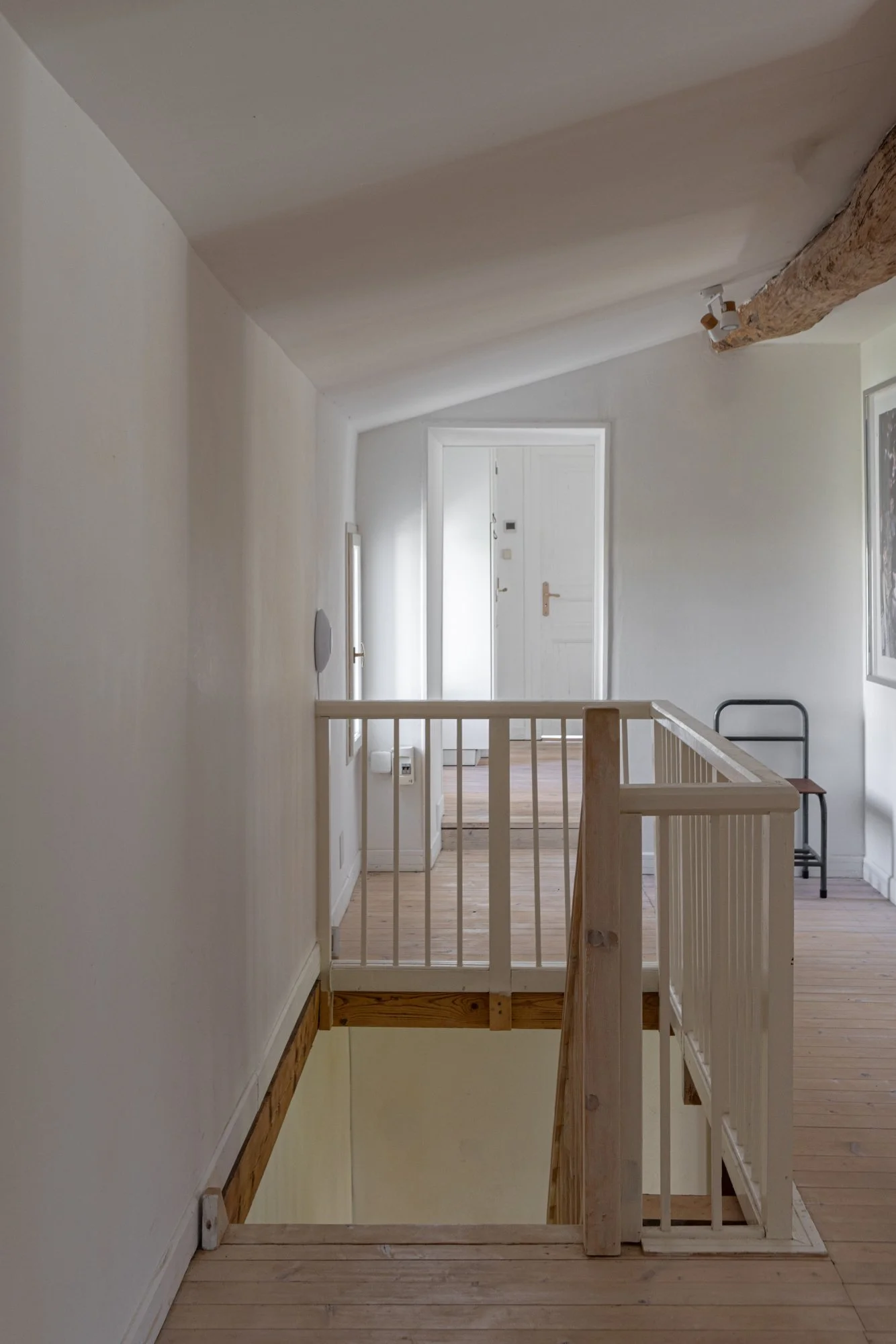 Interior of a house with a staircase, white walls, wooden flooring, exposed wooden beam on the ceiling, and a chair near a framed artwork.