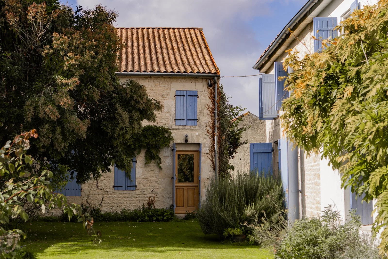 A quaint stone house with blue shutters and a wooden door, surrounded by greenery and a well-manicured lawn.