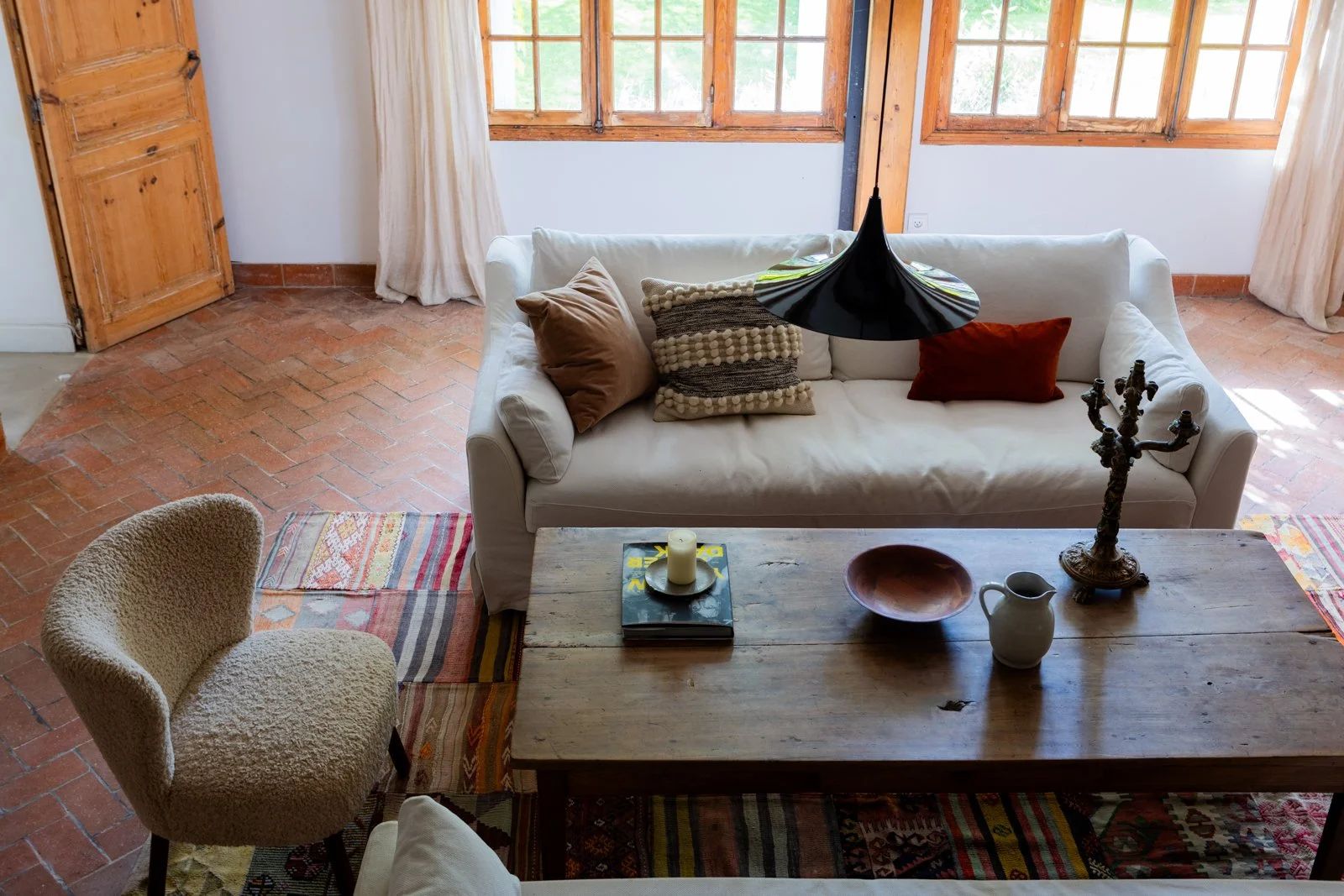 Living room with a white sofa, beige armchair, wooden coffee table, and windows with wooden frames. The room has brick flooring and colorful rugs.