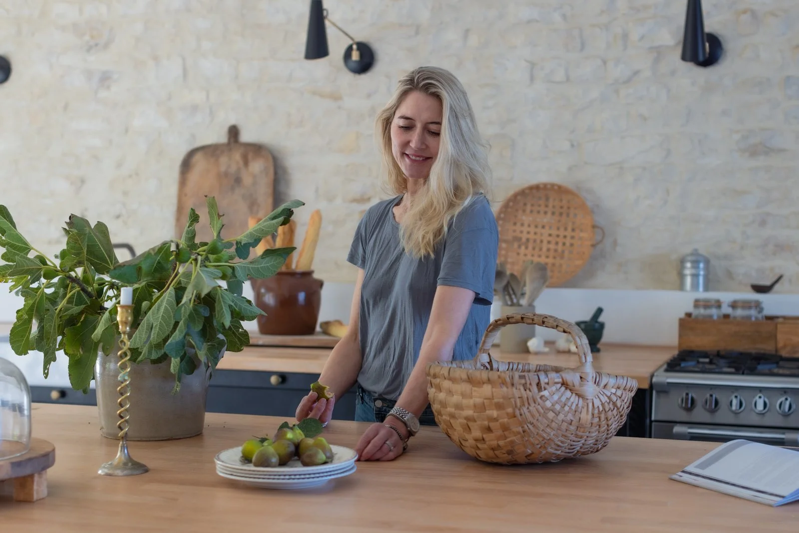 A woman in a gray t-shirt stands in a kitchen with a wooden counter, holding a fig. There is a potted fig tree, a basket, and plates of figs on the counter. The background features a brick wall, hanging black sconces, and kitchen utensils.
