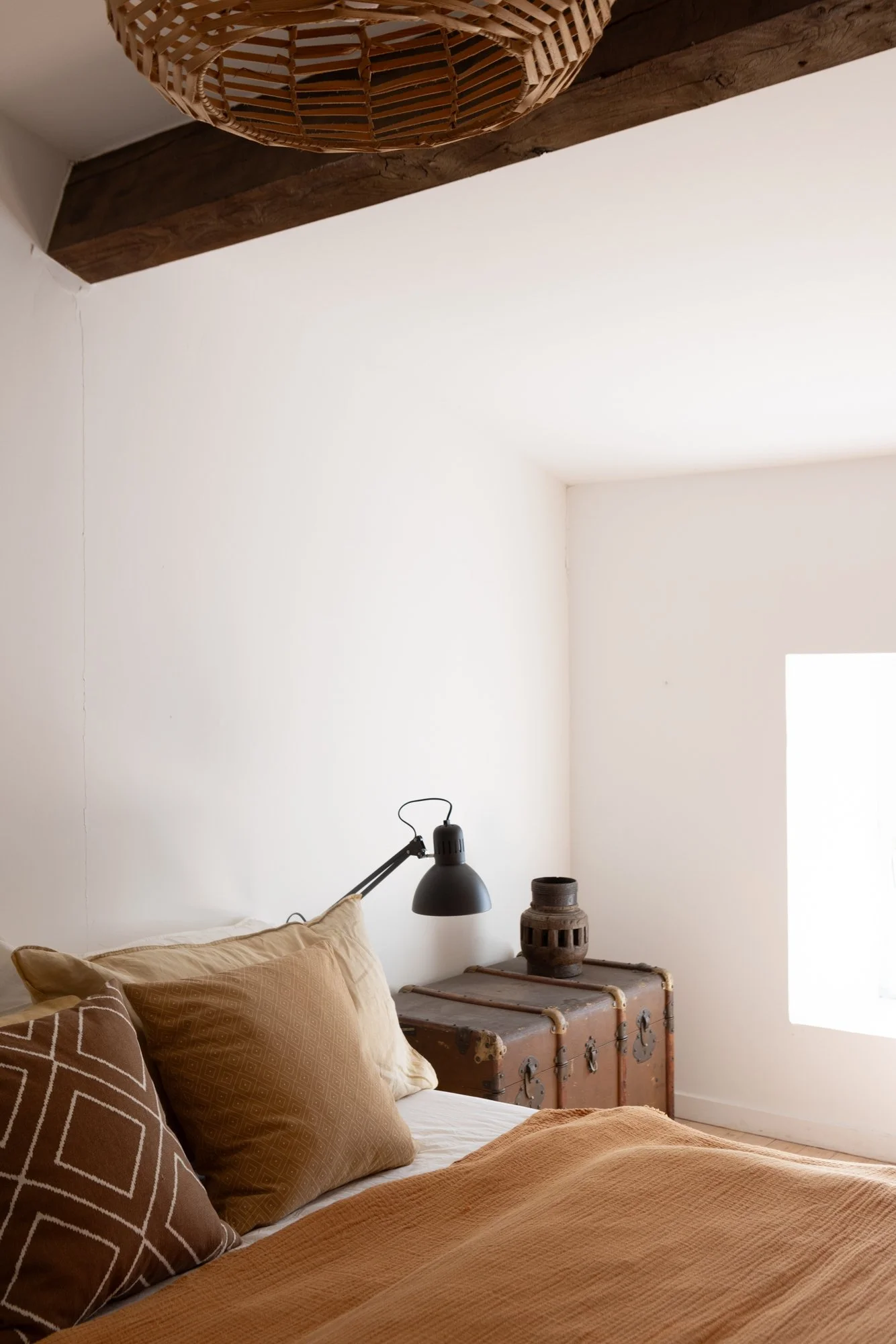 A cozy bedroom with beige pillows, a brown blanket, a black adjustable desk lamp, an antique trunk, and a decorative pottery piece. Natural light from a window illuminates the space with white walls and a wooden beam ceiling.