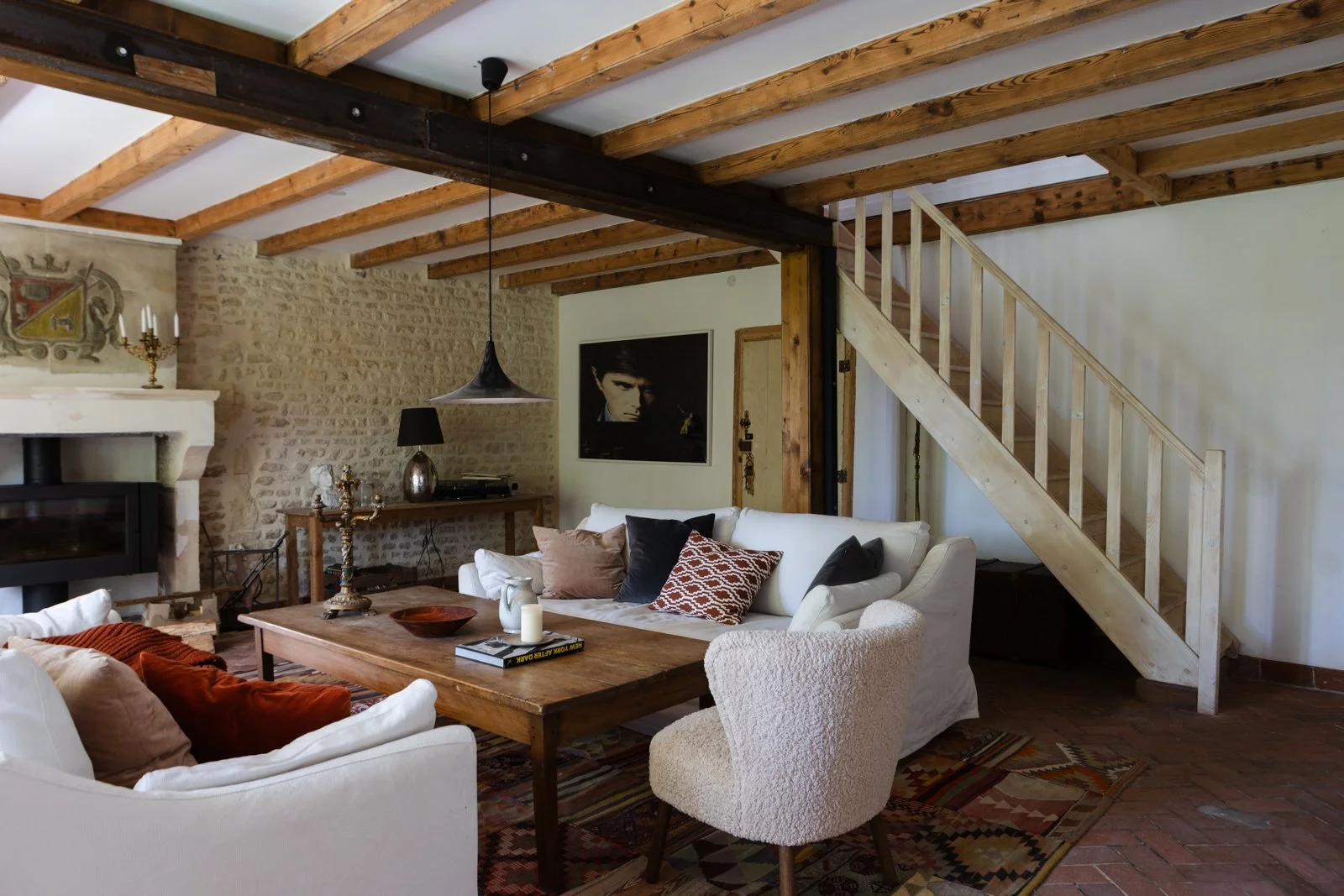 Cozy living room with stone fireplace, wooden beams, white sofas, and a staircase leading upstairs.