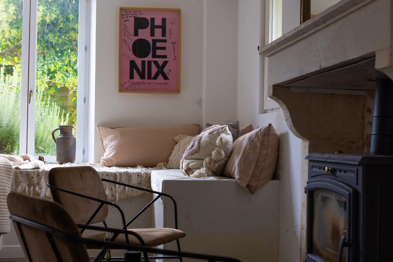 Cozy living room corner with beige cushions and decorative pillows on a built-in window seat, a pink framed 'PHOENIX' poster on the wall, a ceramic pitcher on the windowsill, and a black wood stove near a stone fireplace with sunshine streaming throu