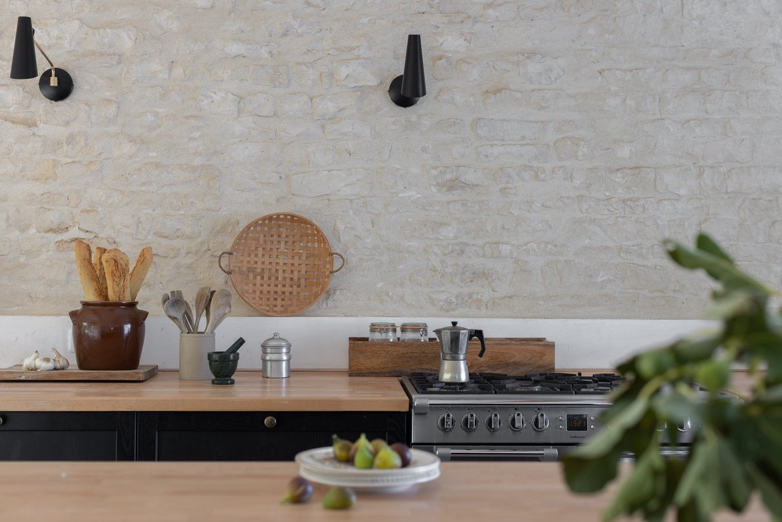 Kitchen with light-colored brick wall, wooden countertop, black stove, and various kitchen items including a pot with bread, garlic, utensils, a decorative tray, a moka pot on the stove, a plate of figs, and green leaves in the foreground.
