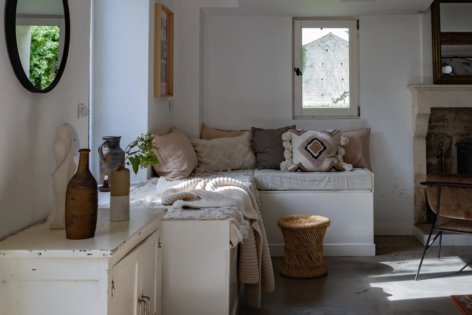 Sunlit cozy living room with a white built-in sofa, decorated with pillows and a chunky knit blanket. There are decorative vases and a small plant on a white side table. A wicker stool is placed in front of the sofa. The room features a window with a