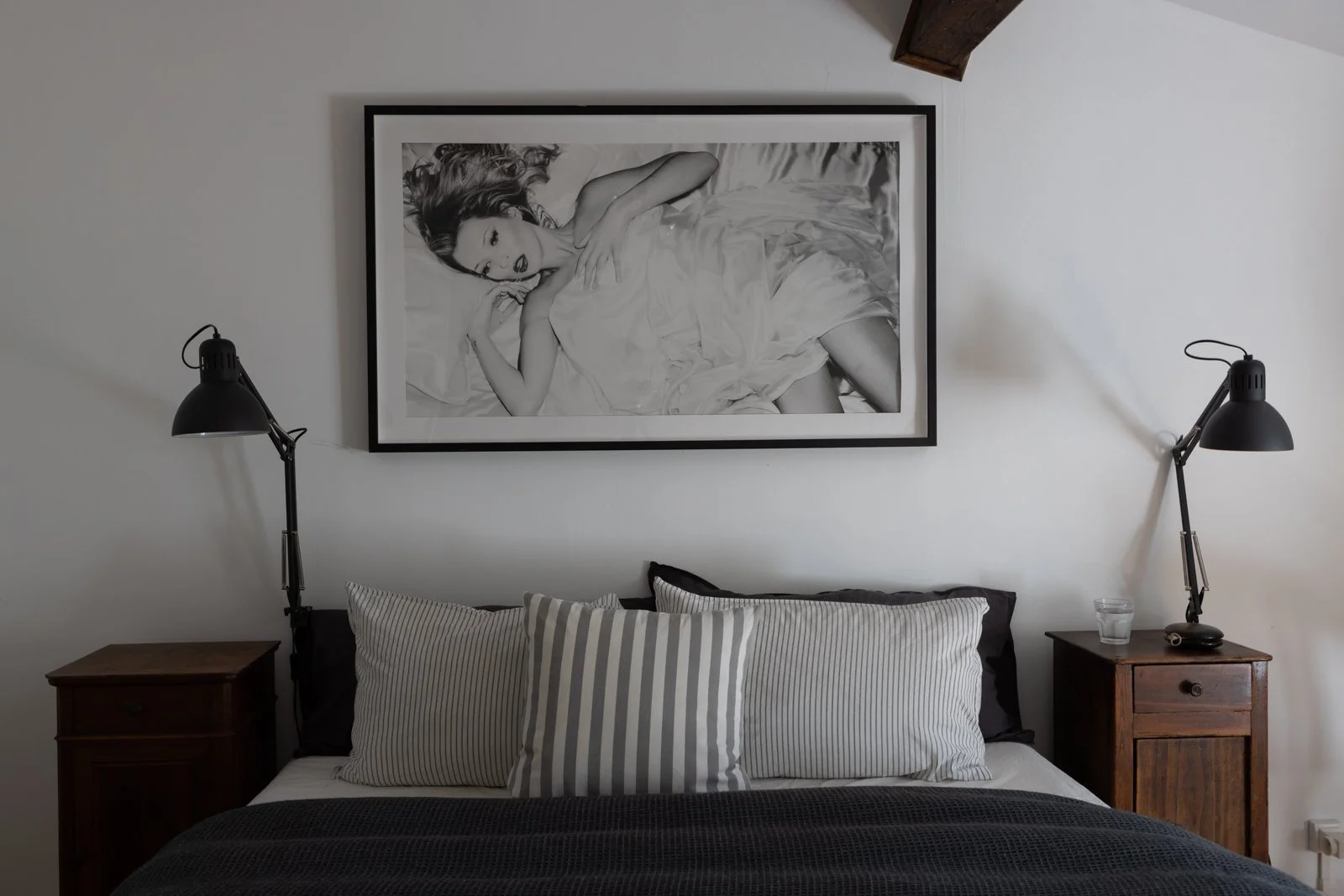 A bedroom with a bed, three striped pillows, two black pillows, and a black bedspread. There are two wooden nightstands with black adjustable lamps, one with a glass of water and a remote. A large black and white framed photograph of a woman lying in