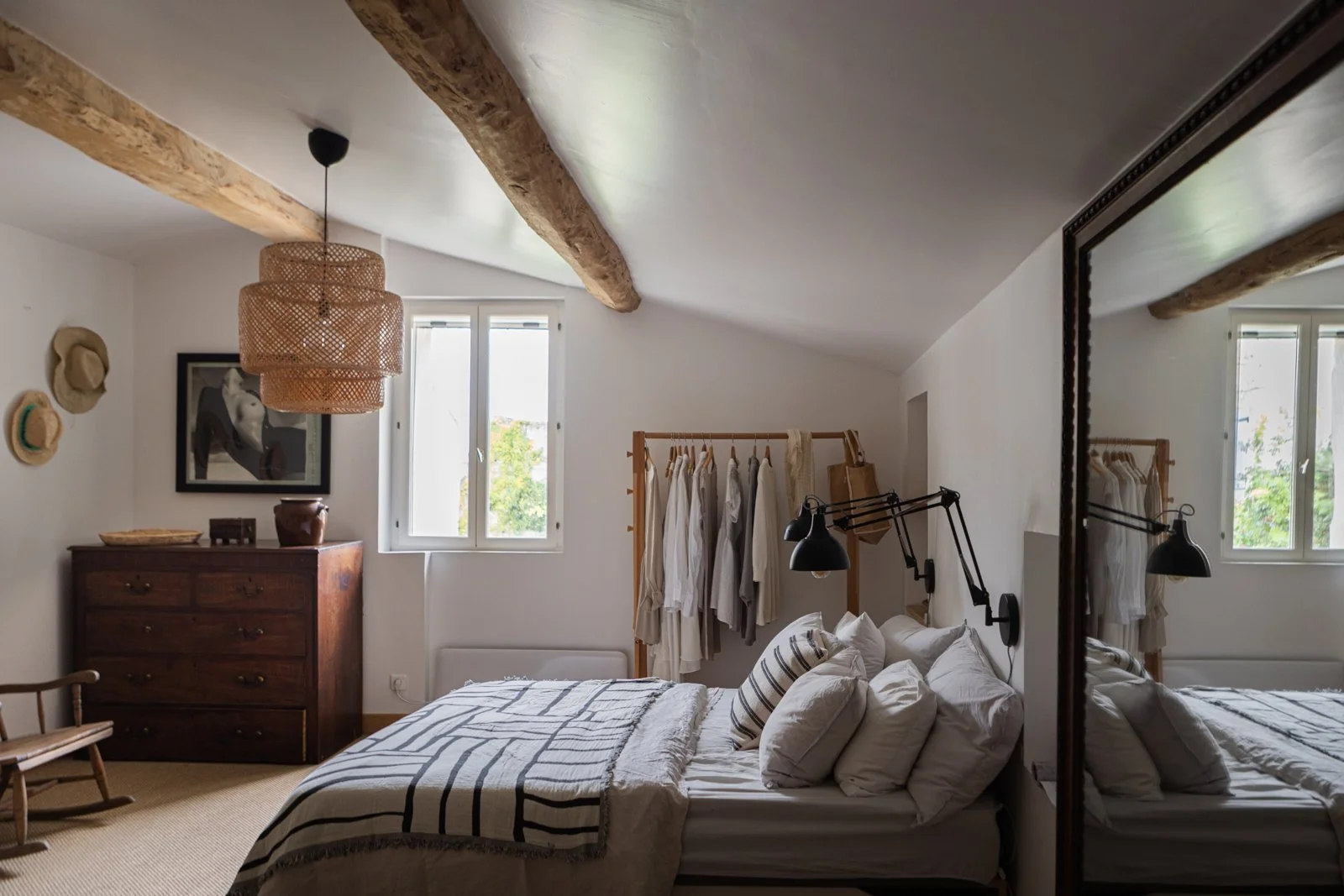 A bedroom with white walls, a sloped ceiling, and exposed wooden beams. It has a bed with white linens and black accents, a wooden dresser, a hanging rack with clothes, a mirror, and a window with a view of green trees outside.