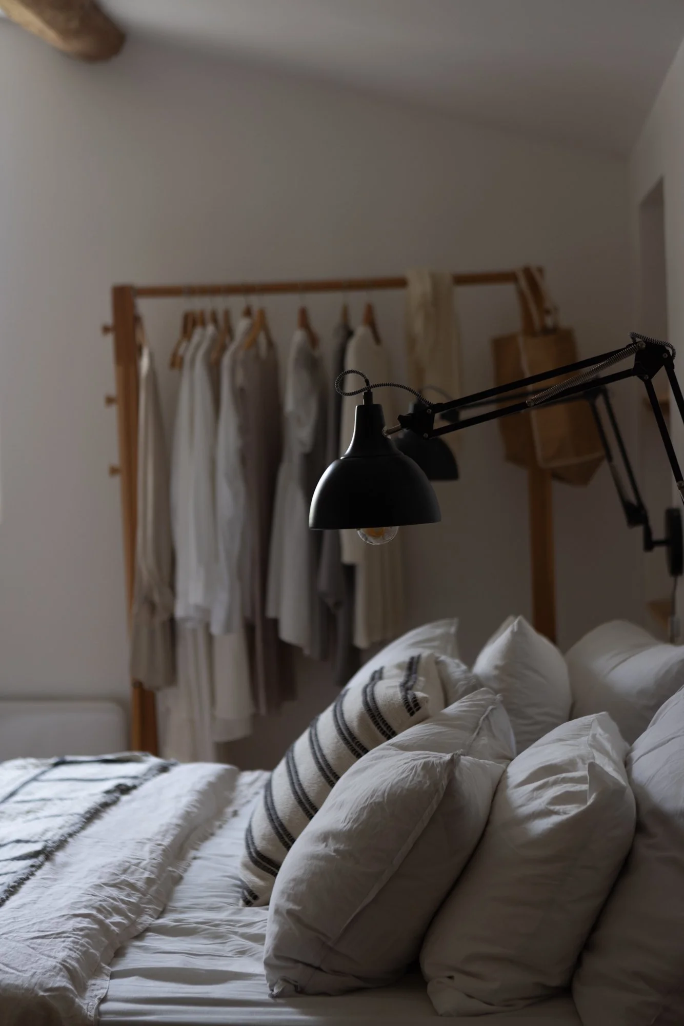 A bed with multiple white pillows, a blanket, and a striped cushion, in a bedroom with a black adjustable wall lamp. In the background, there is a clothing rack with hanging white and beige dresses and tops.