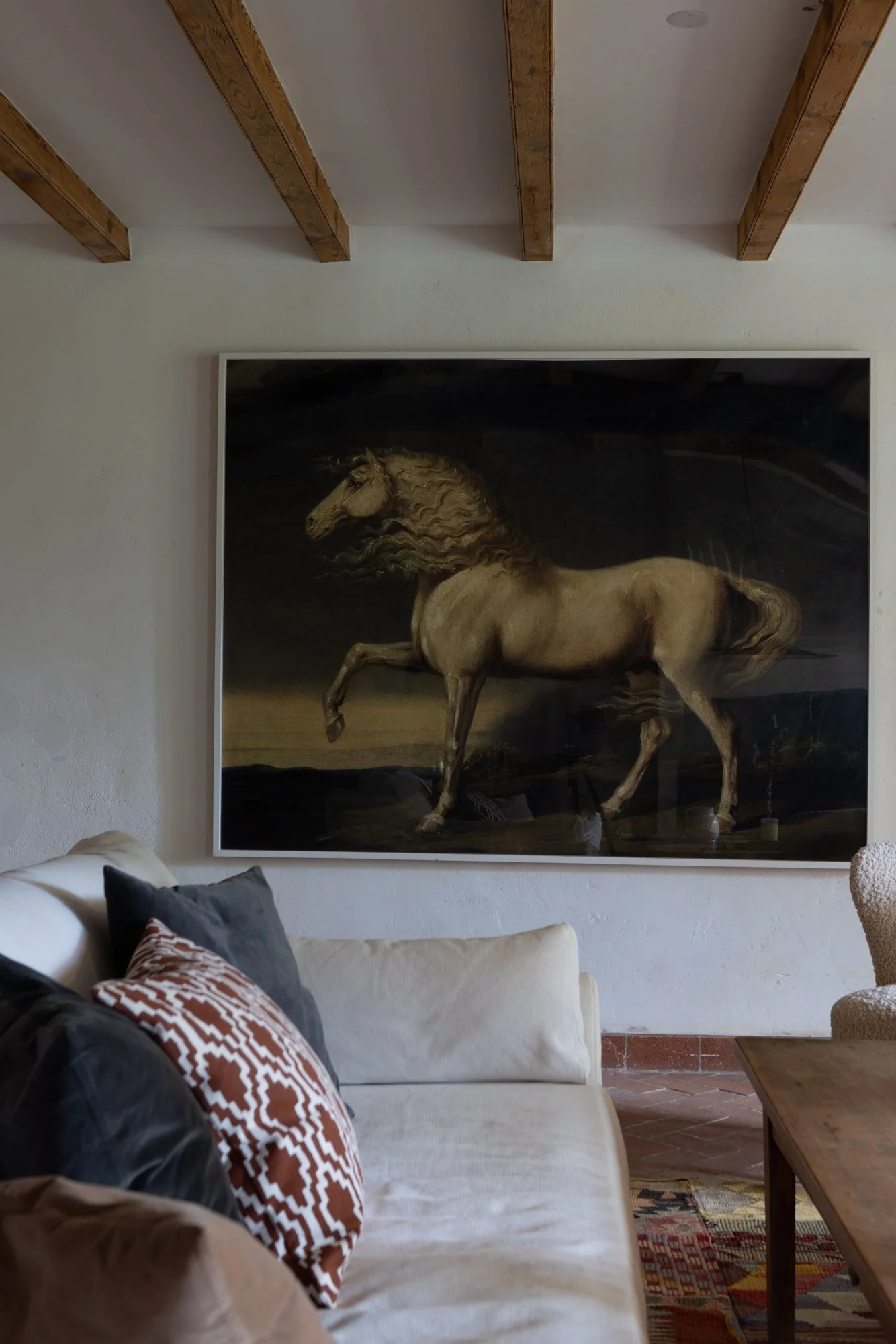 Interior of a room with a beige sofa and decorative pillows, a wooden coffee table, and a large framed painting of a galloping horse on the wall with exposed wooden ceiling beams.