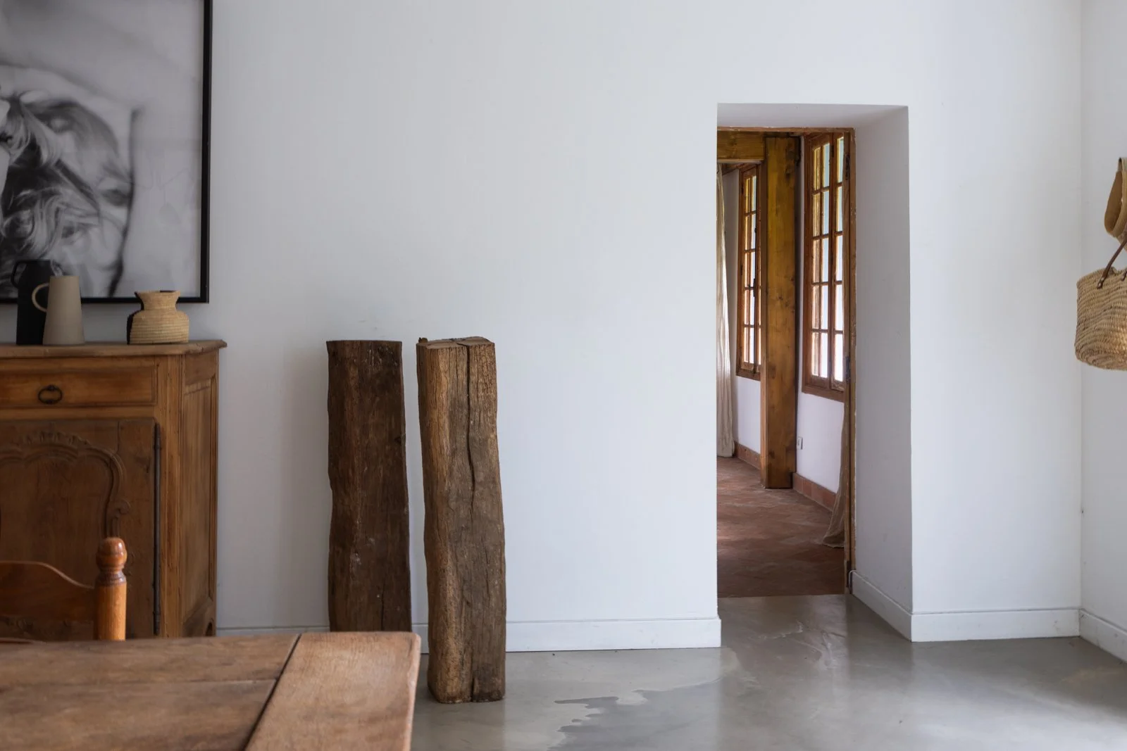 Interior of a room with white walls, wooden furniture, and two vertical wooden logs leaning against the wall. Open door reveals another room with wooden window frames and a partially visible curtain.