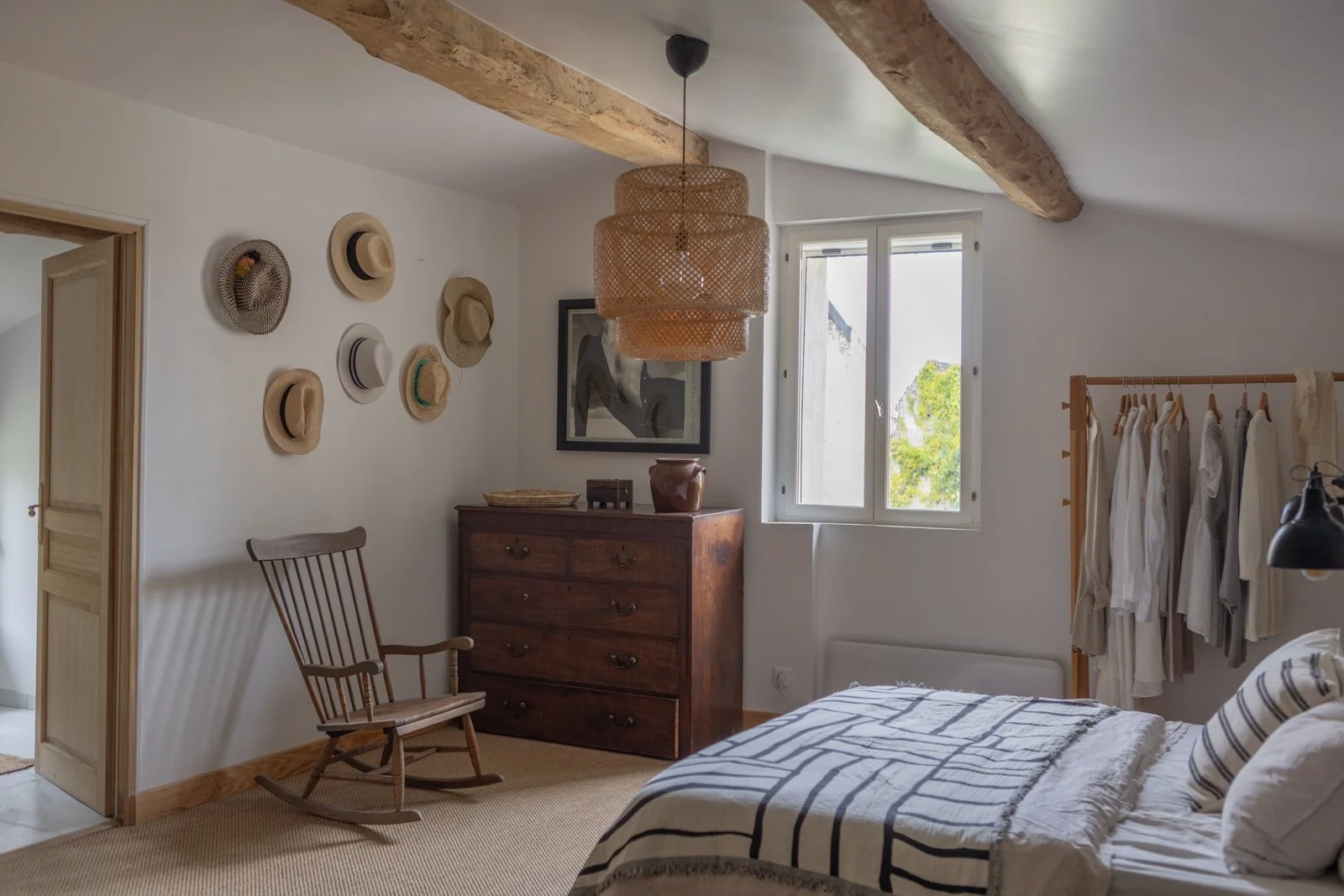 A cozy bedroom with white walls, a wooden dresser, a rocking chair, a bed with striped bedding, and a clothing rack with white and beige clothes. Natural light coming through a window, with a decorative wall of hats and a framed abstract picture.