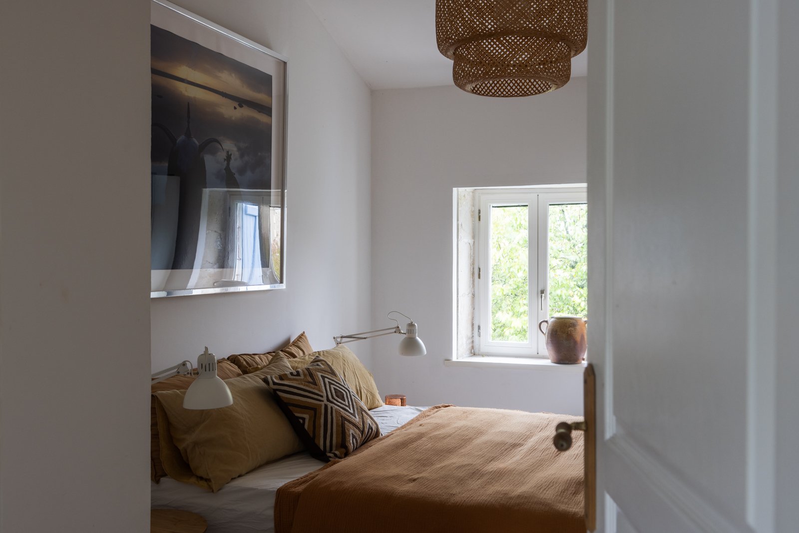 A cozy bedroom with white walls, a bed with beige and brown pillows, and an orange blanket. A small window with a view of green trees outside. A large framed picture hangs on the wall above the bed. A ceiling light fixture and wall-mounted lamps are 