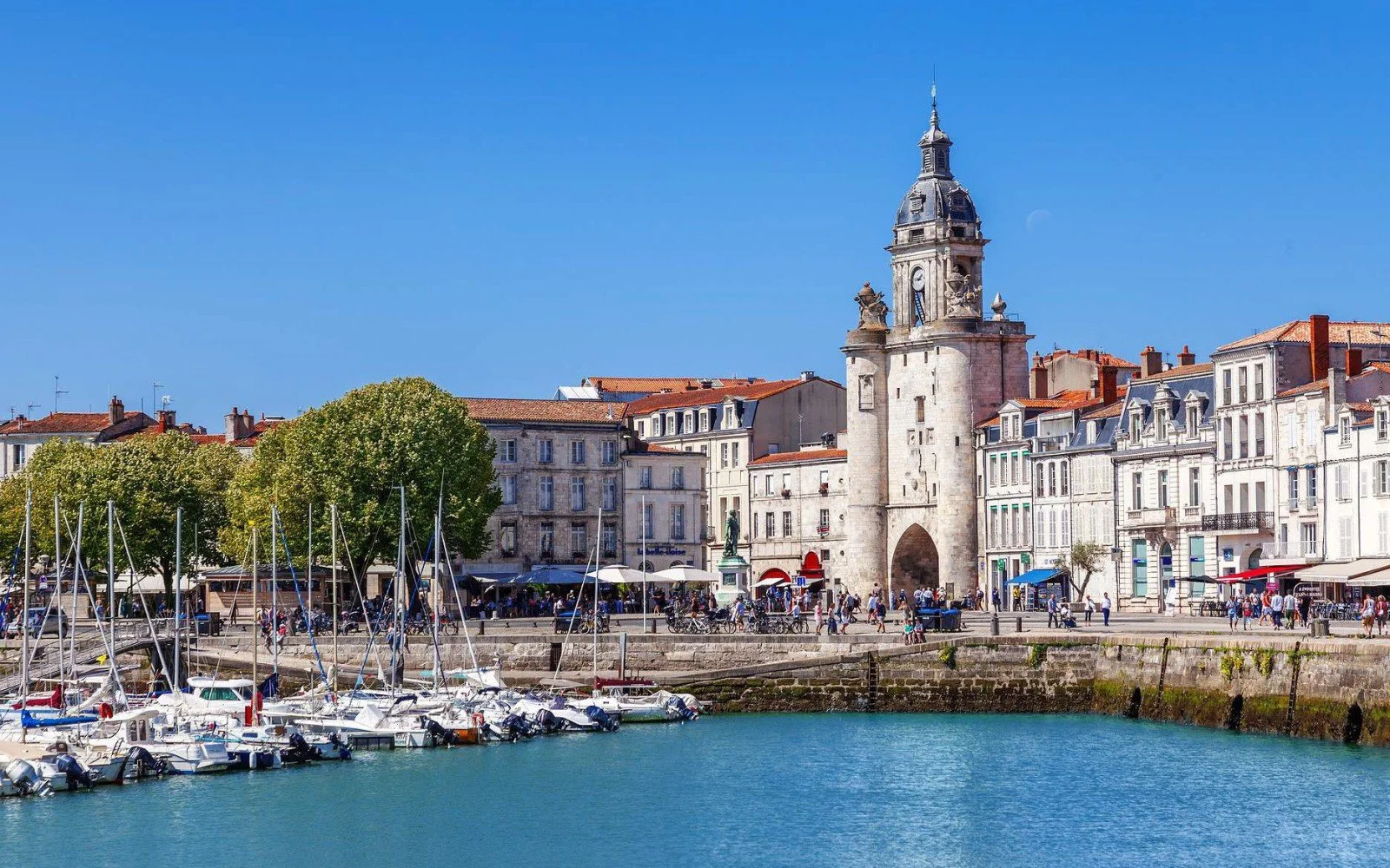 A city waterfront with boats docked at a marina, historic buildings with red roofs, and a prominent clock tower, under a clear blue sky.
