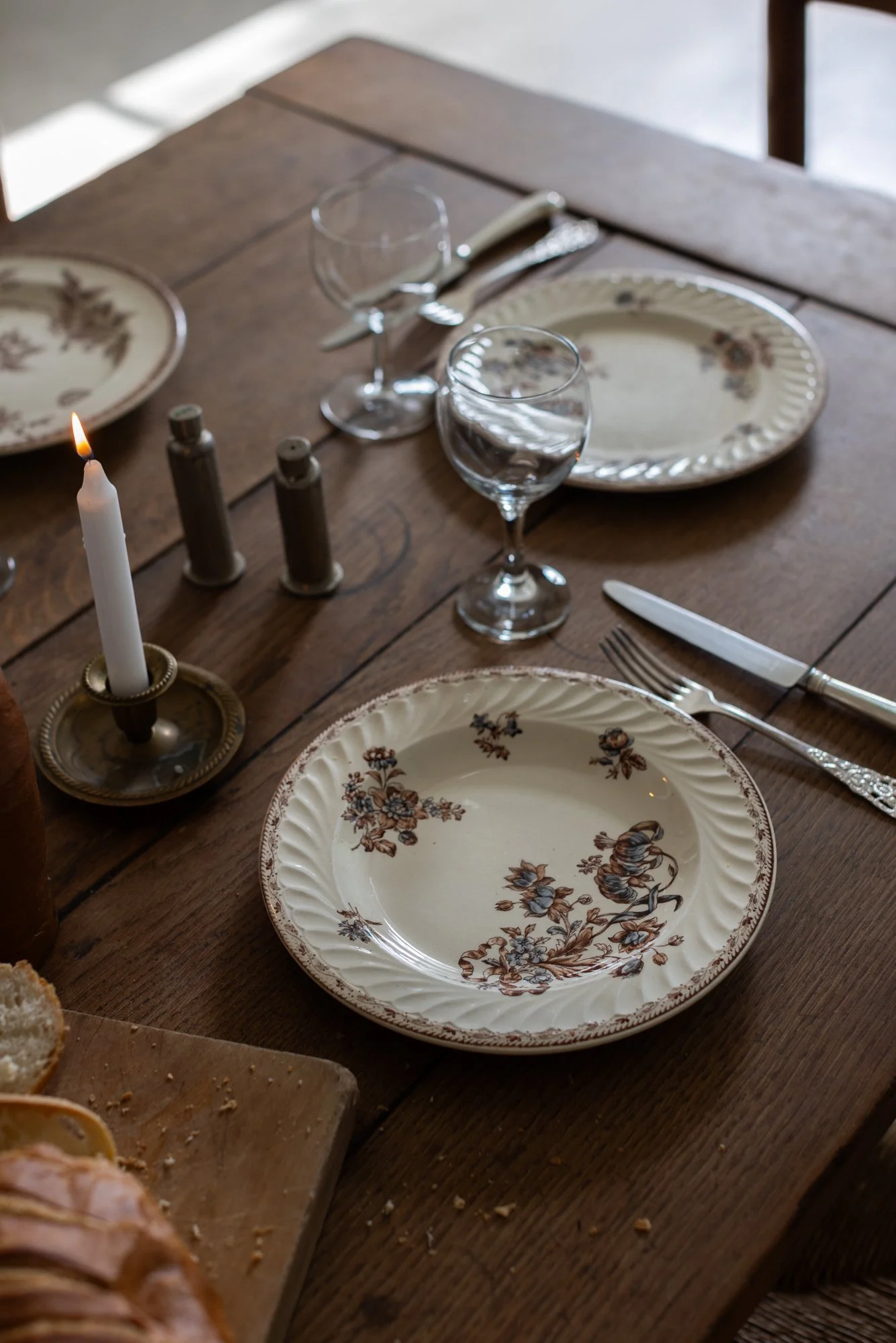 Wooden dining table set with floral-patterned plates, wine glasses, forks, knives, salt and pepper shakers, and a lit candle in a brass holder.