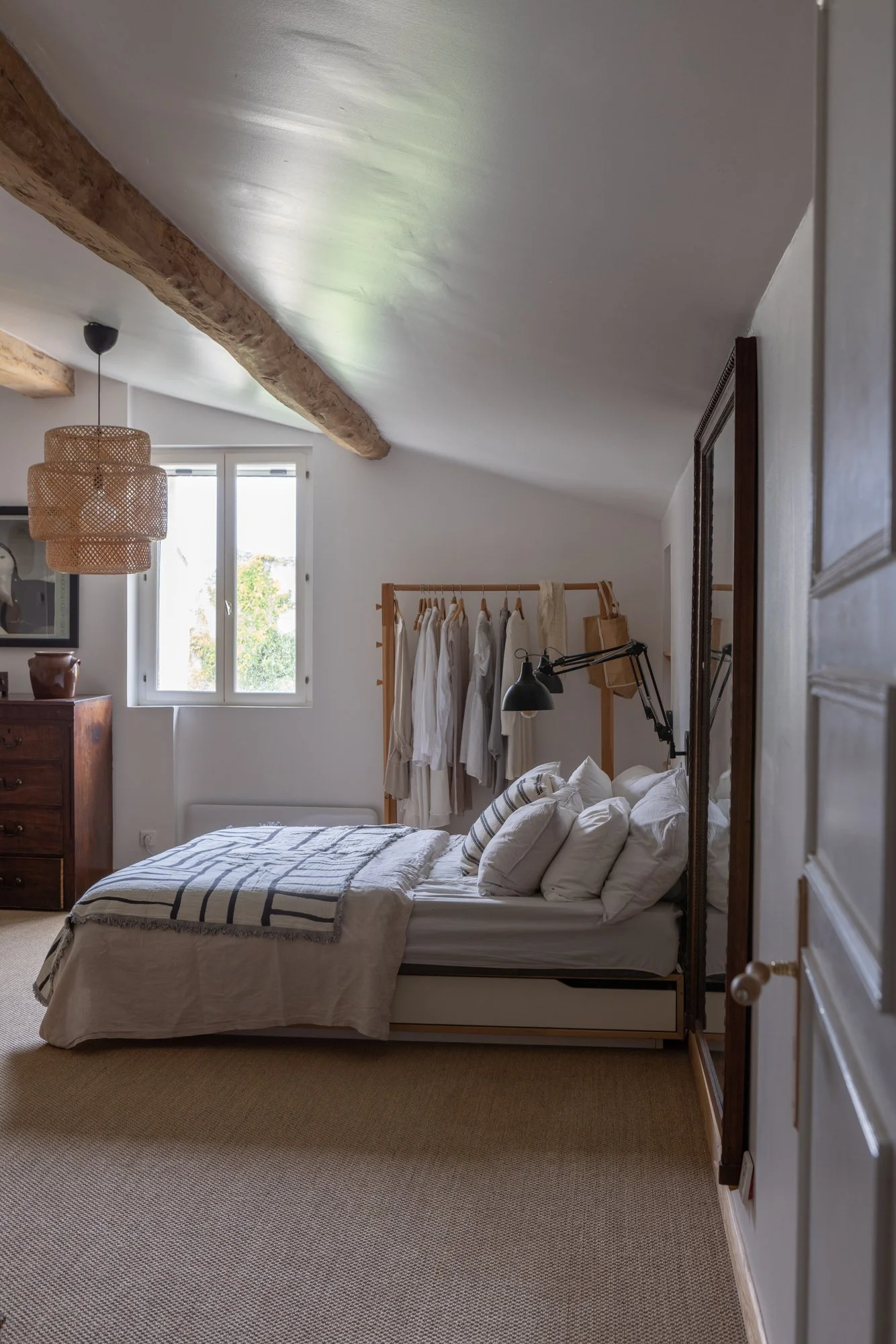 A cozy bedroom with a sloped ceiling, white walls, and natural light coming through a window. It features a bed with white bedding and multiple pillows, a wooden in-room clothing rack with hanging clothes, a black adjustable wall lamp, a wooden dress